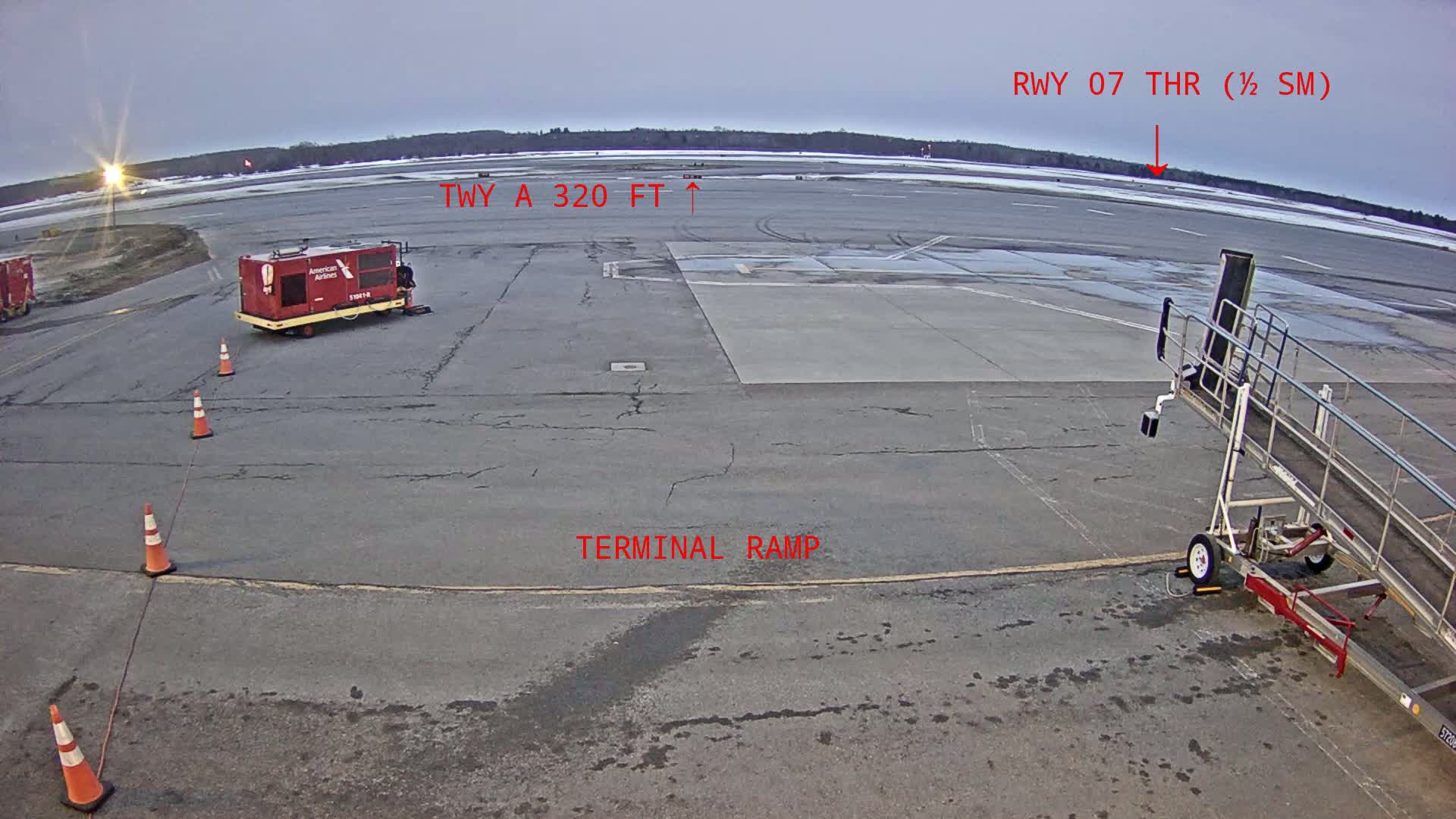 An airport tarmac under an overcast sky shows a jetway, ground support equipment, and orange traffic cones near a terminal ramp.