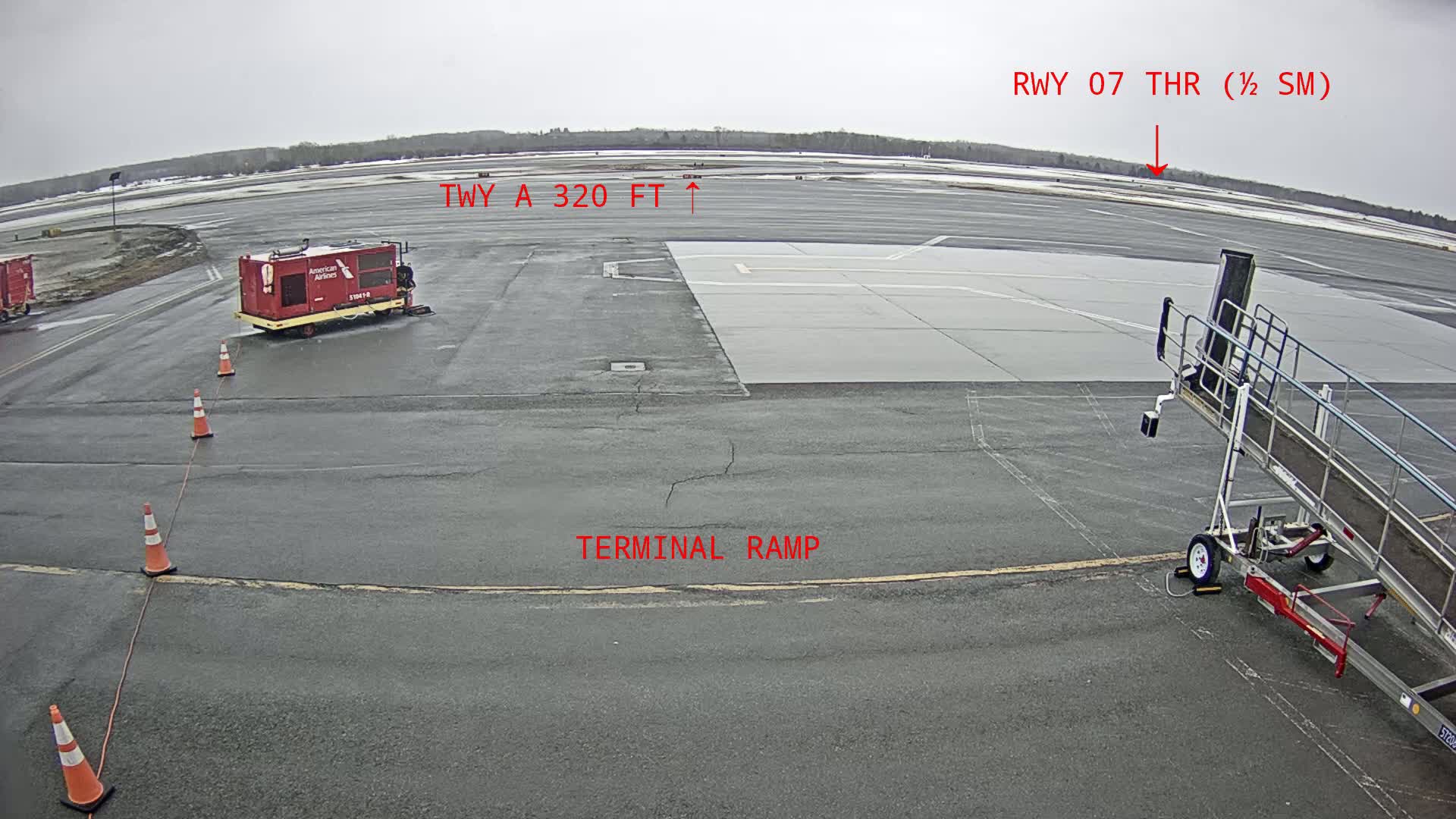 A tarmac with a jet bridge, a baggage cart, and traffic cones, under an overcast sky.