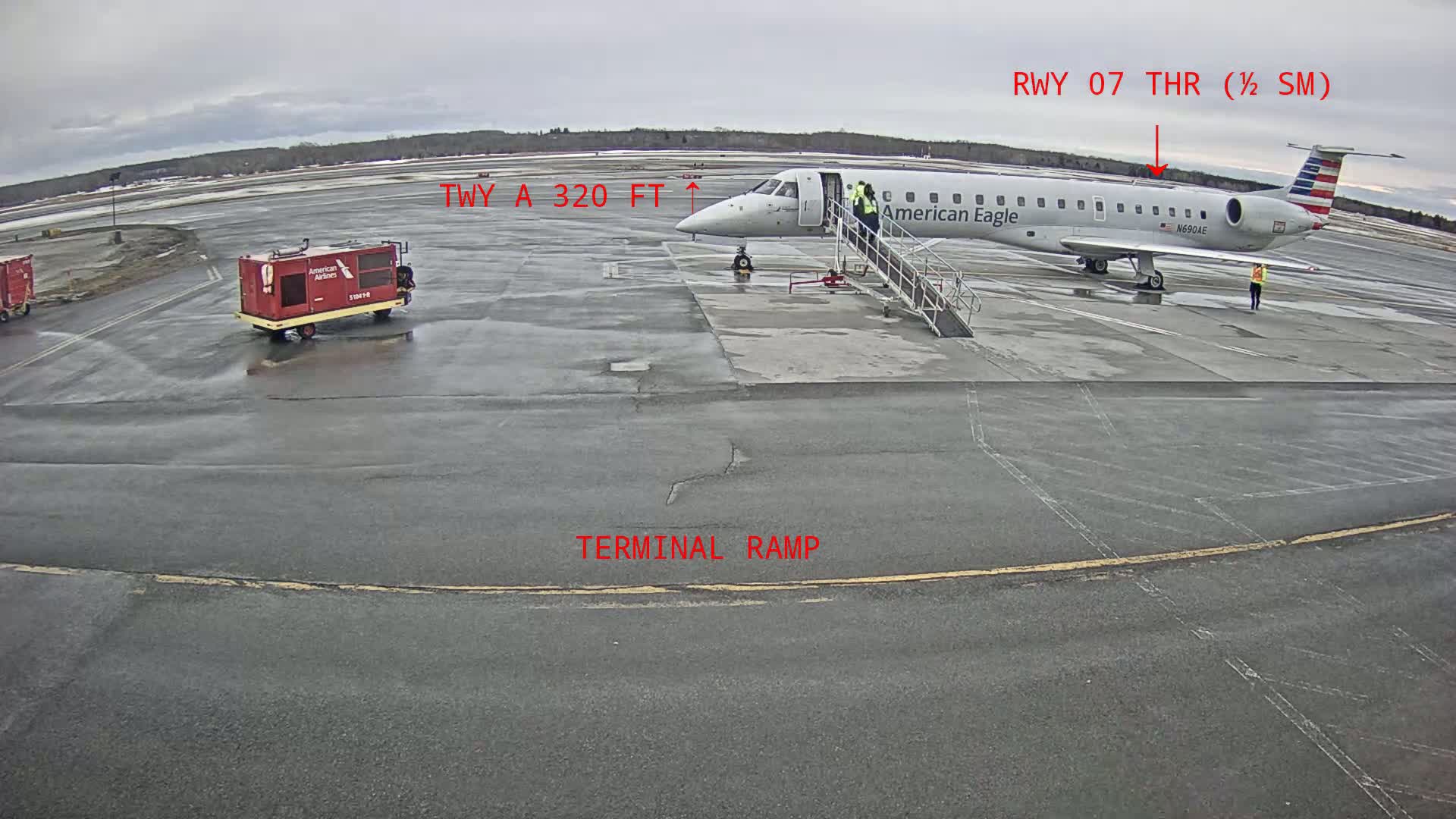 An American Eagle airplane is parked at a terminal gate on a wet tarmac, with ground support equipment nearby under an overcast sky.