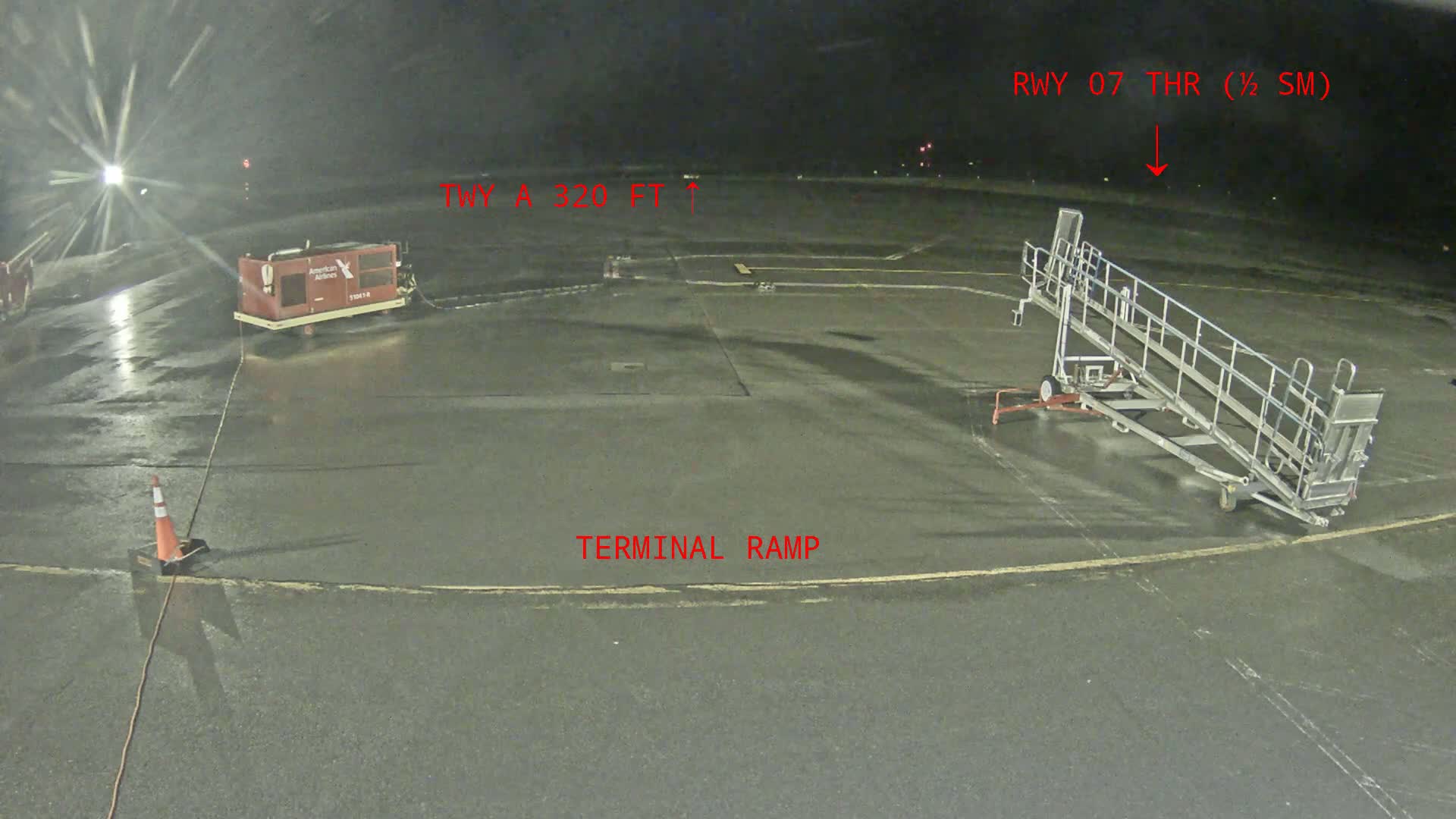 A wet airport tarmac at night shows a baggage cart, a passenger boarding ramp, and a single traffic cone.