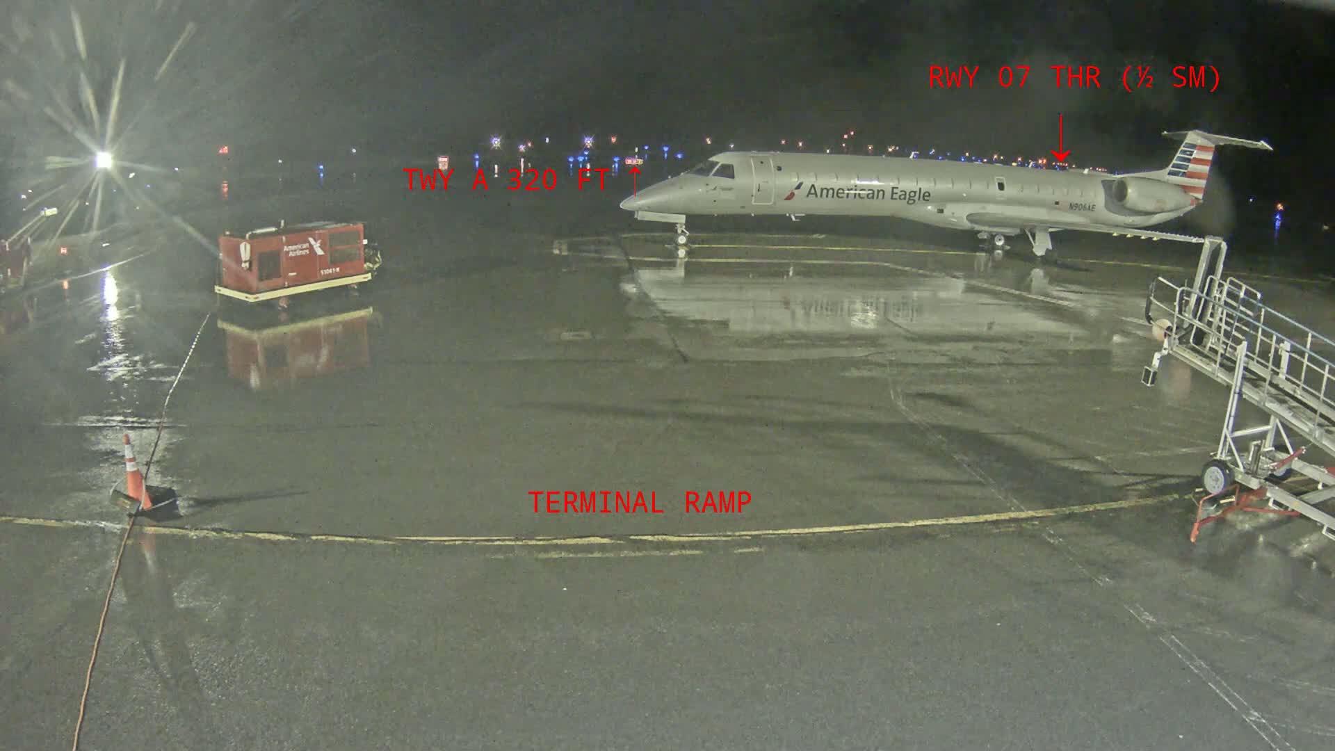 At night, under rainy conditions, an American Eagle plane sits on a wet tarmac near a jet bridge and ground support equipment.