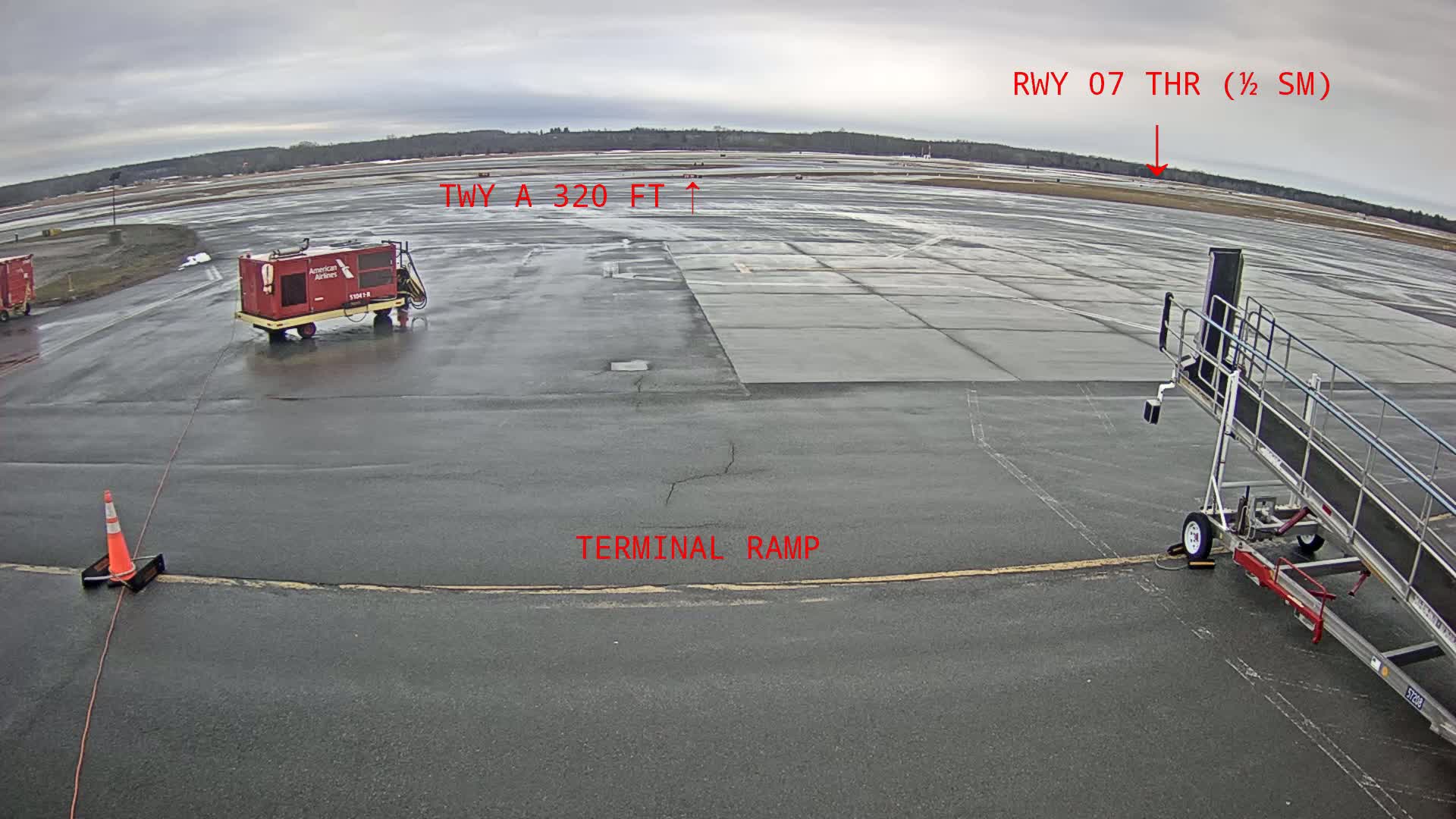 A wet airport tarmac is shown, with an airplane servicing vehicle and a passenger boarding bridge near a terminal ramp, under an overcast sky.