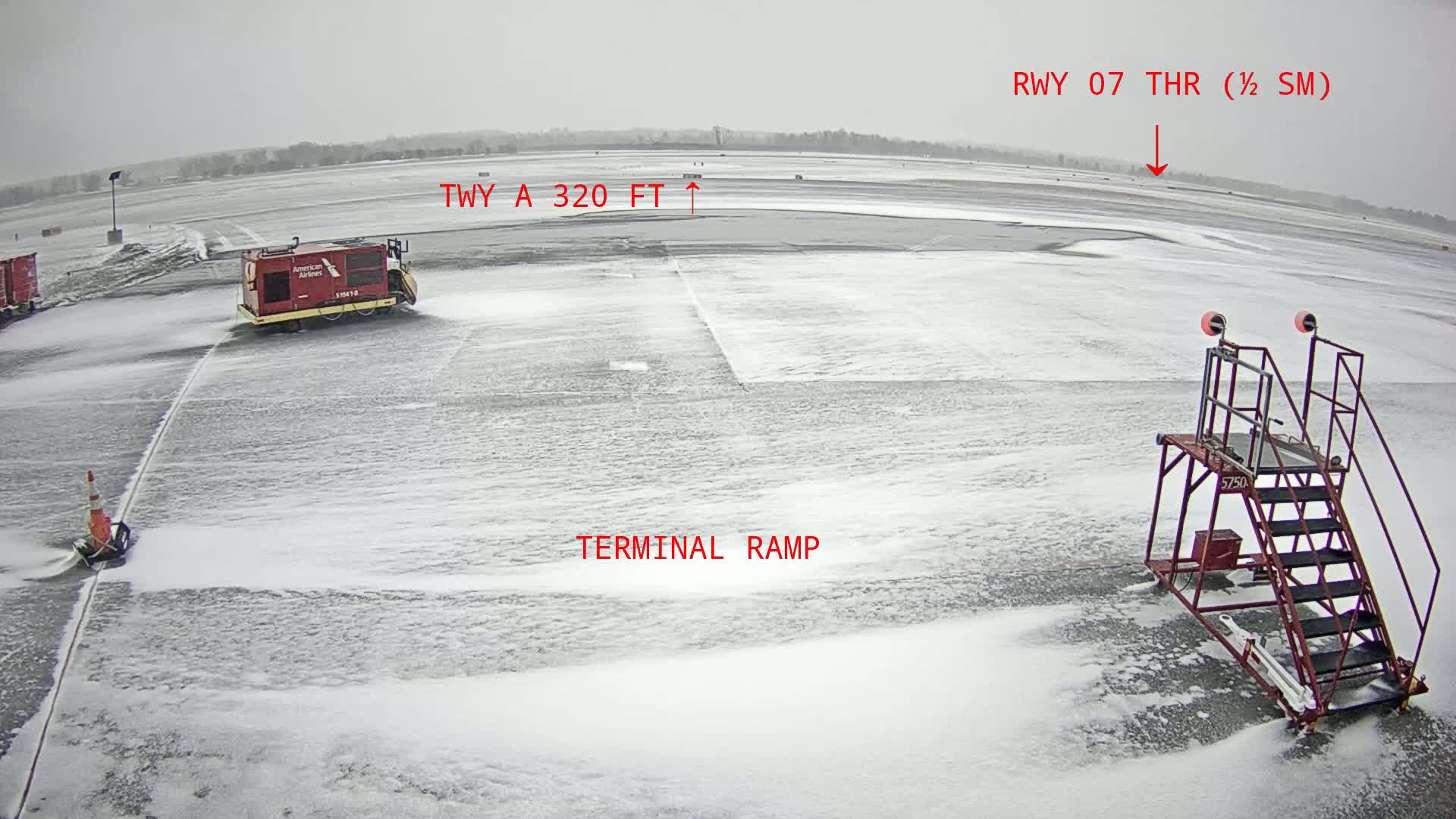 A snow-covered airport tarmac shows a de-icing vehicle and airport equipment under a heavy snowfall.