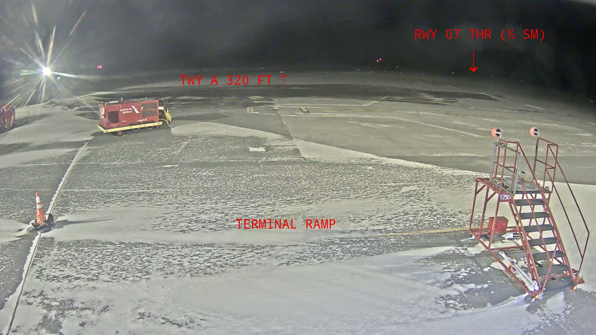 A snow-covered airport tarmac at night shows a baggage tug and a stair unit near a terminal ramp, with runway lights visible in the distance.