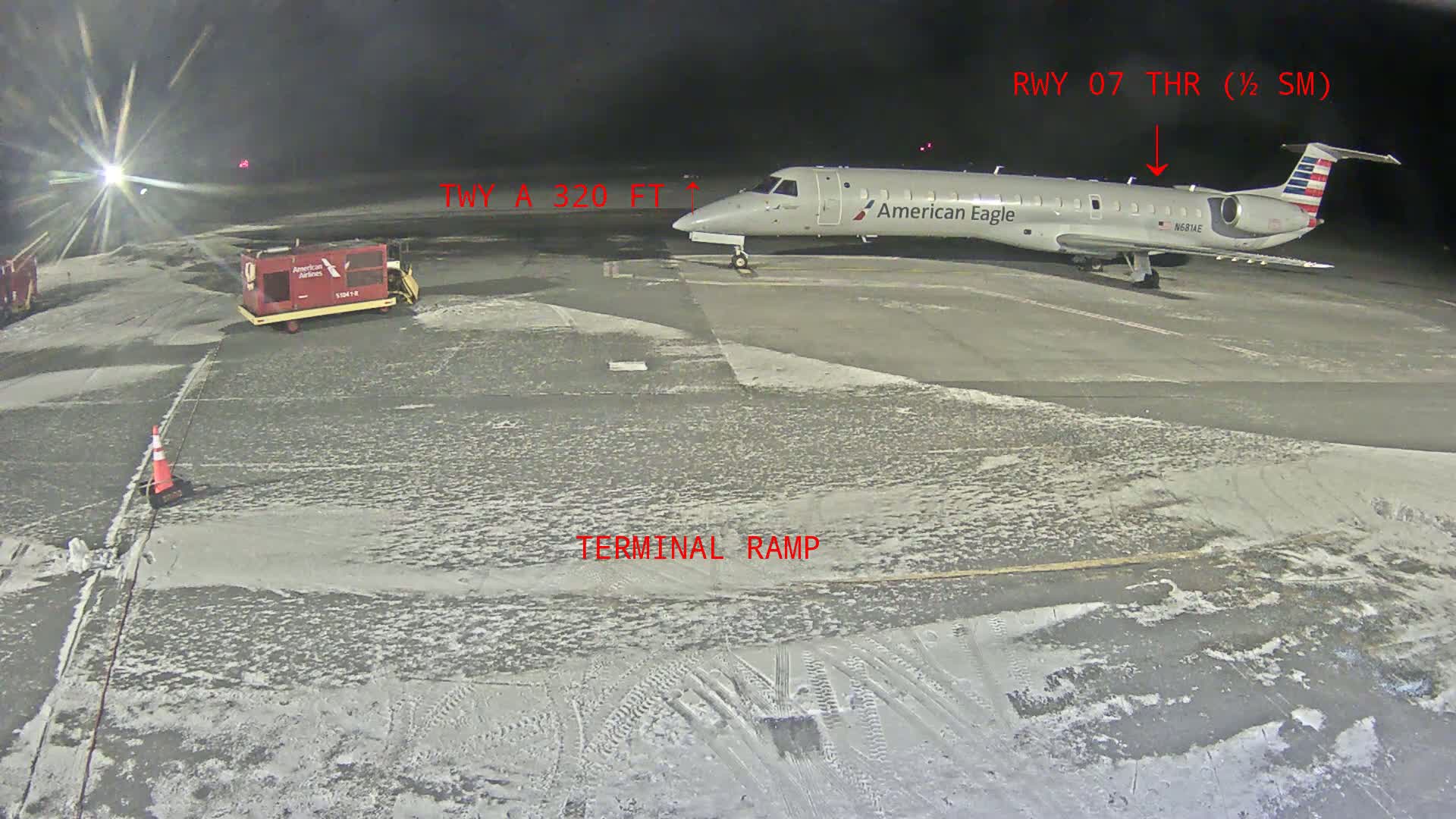 An American Eagle regional jet is parked on a snow-covered tarmac at night, near a baggage tug.