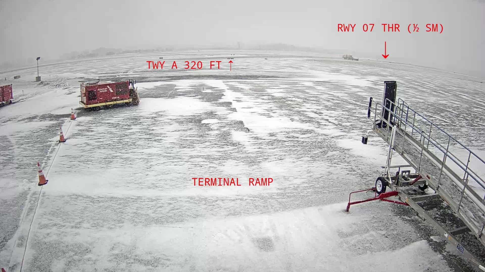 A snow-covered airport tarmac shows a jet bridge, a snow removal vehicle, and orange traffic cones during a heavy snowfall.