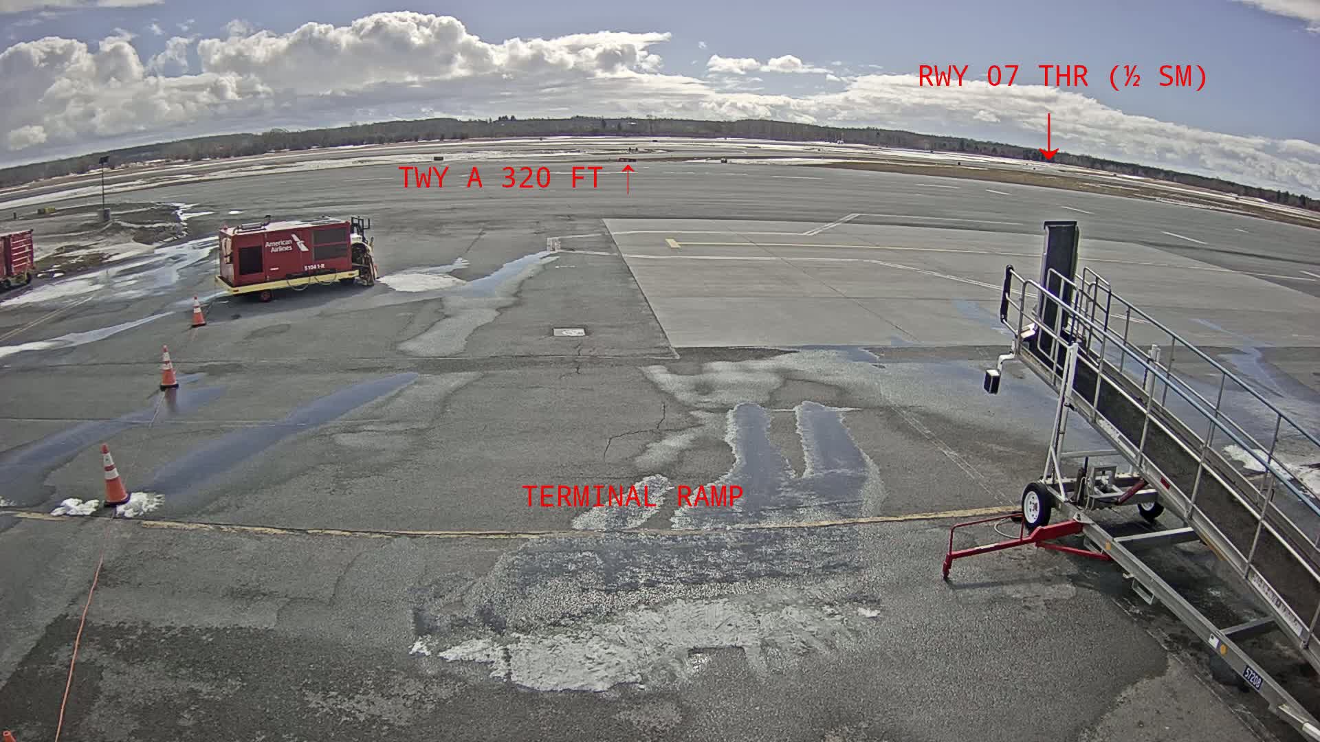 A mostly sunny day at an airport tarmac shows a jet bridge, service vehicles, and patches of melting snow or ice on the pavement.