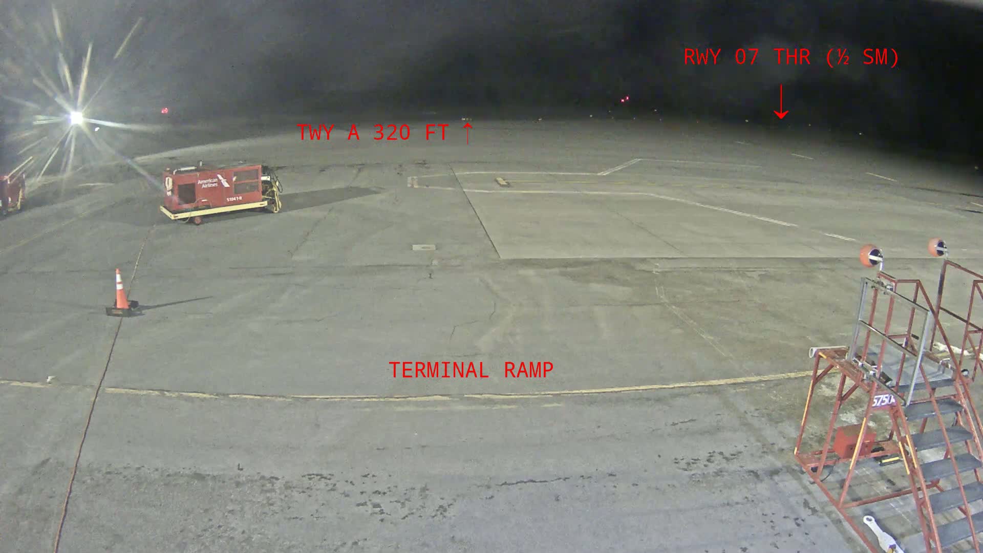 A nighttime view of an airport tarmac shows a baggage cart, a traffic cone, and airport equipment under dark, clear skies.