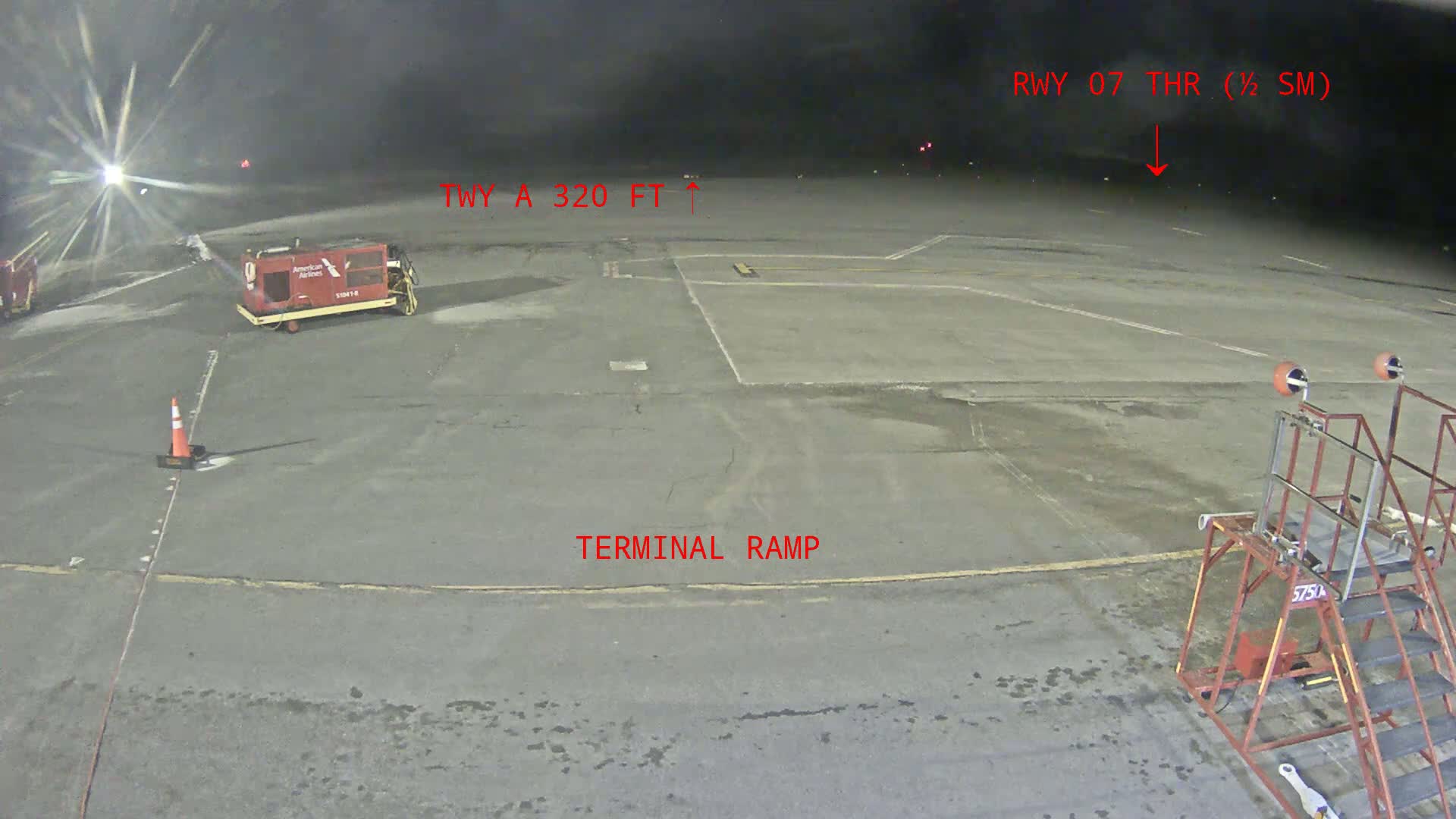 A nighttime view of an airport tarmac shows a baggage tug, a terminal ramp, and airport ground support equipment under a dark, possibly cloudy sky.