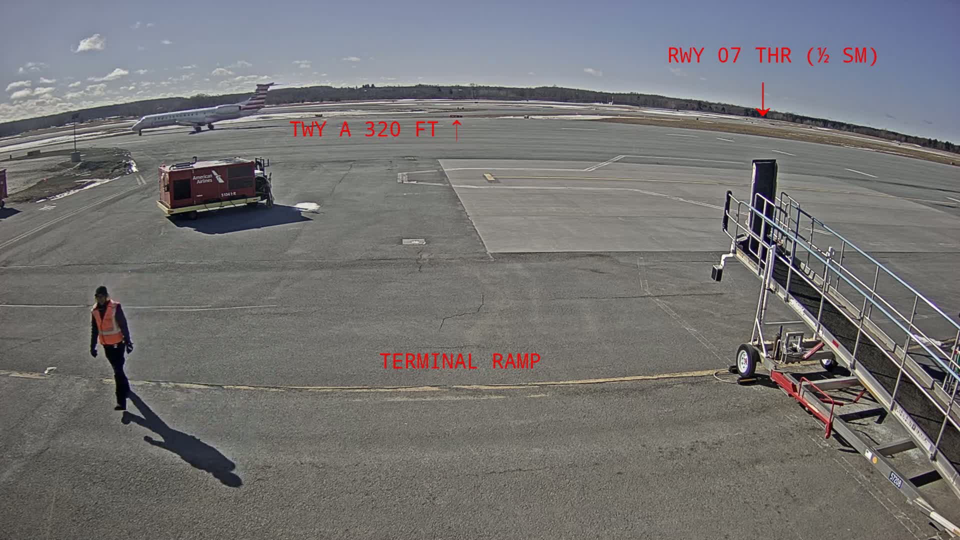 A person in a safety vest walks across an airport tarmac on a sunny day, near a parked plane, a baggage tug, and a jet bridge.