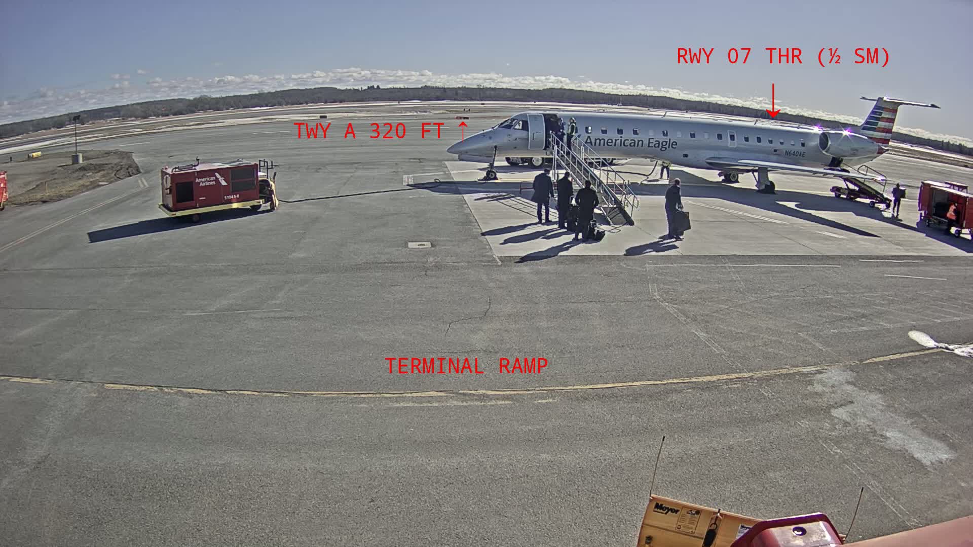 An American Eagle regional jet is parked at a terminal ramp on a sunny day, with passengers disembarking and ground crew present.