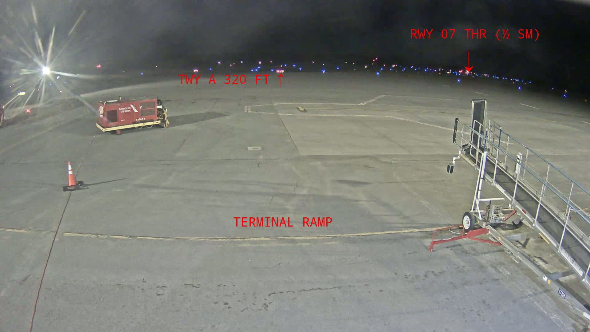 A nighttime view of an airport tarmac shows a terminal ramp, a baggage tug, and a jet bridge, with several aircraft visible in the distance under dark skies.