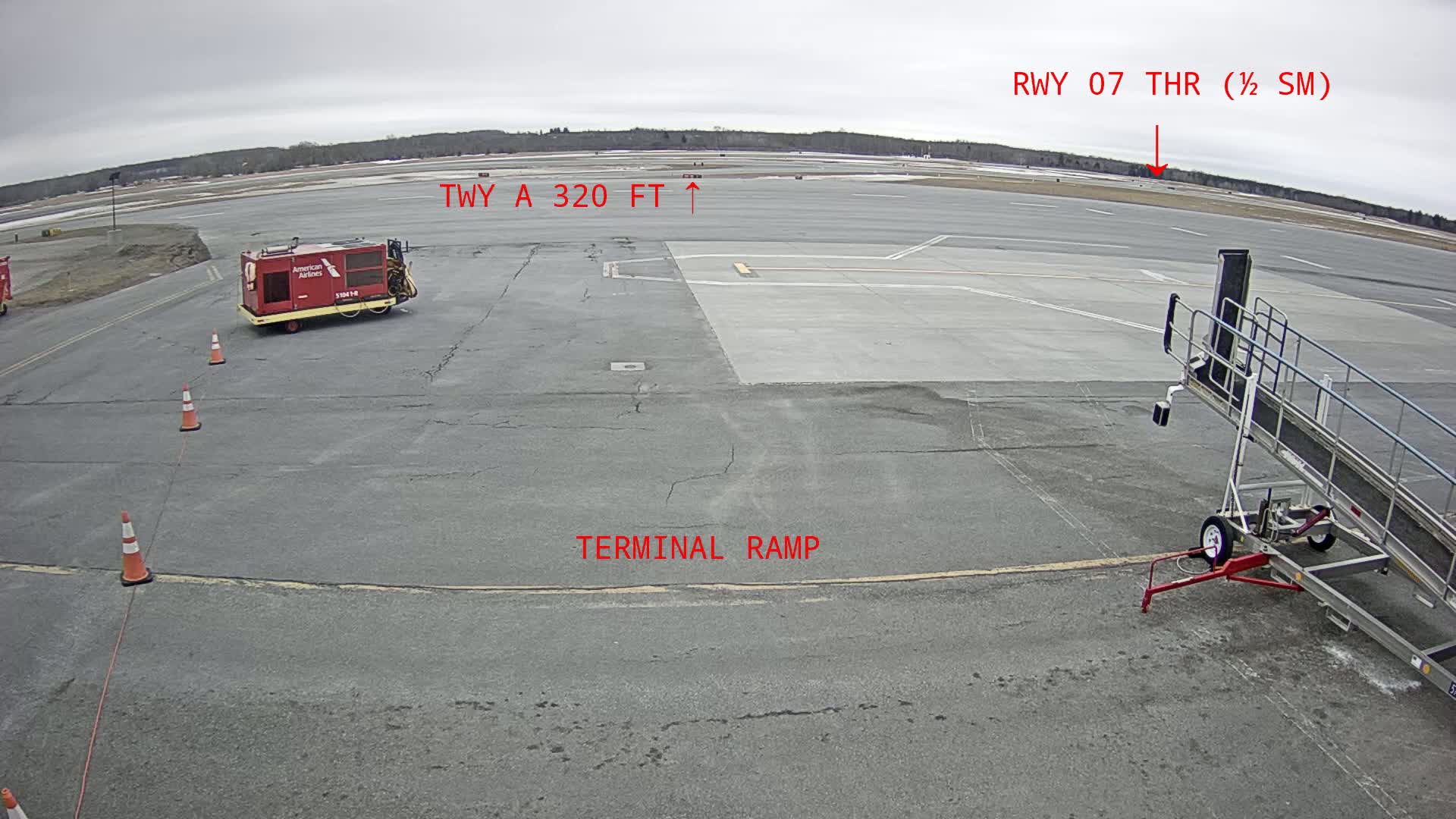 An airport tarmac on an overcast day shows a red baggage cart, an empty jet bridge, and orange traffic cones near a terminal ramp with a runway visible in the distance.
