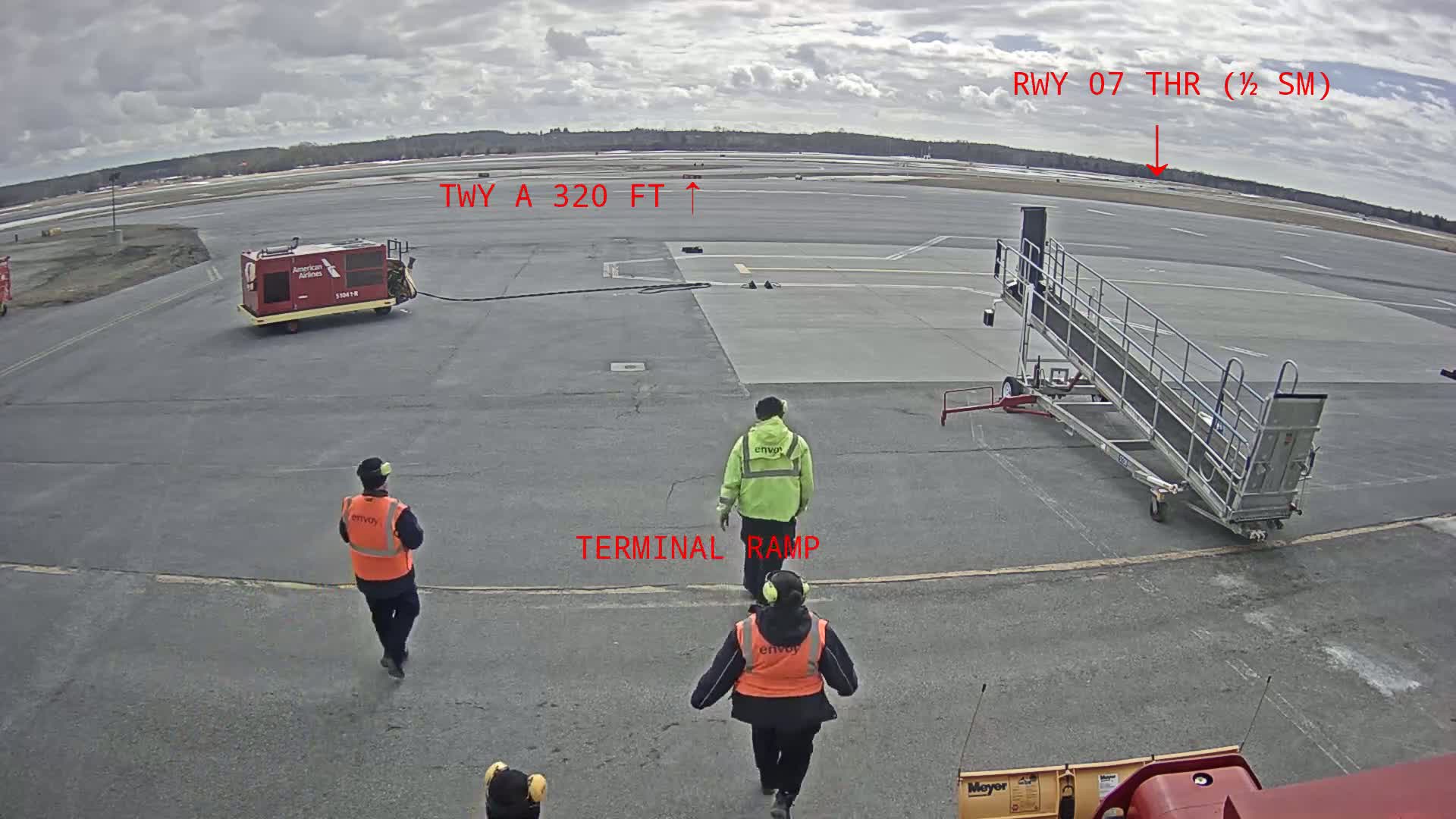 On an overcast day at an airport, three ground crew members walk towards a terminal ramp, past an American Airlines fuel truck and a passenger boarding stairs.