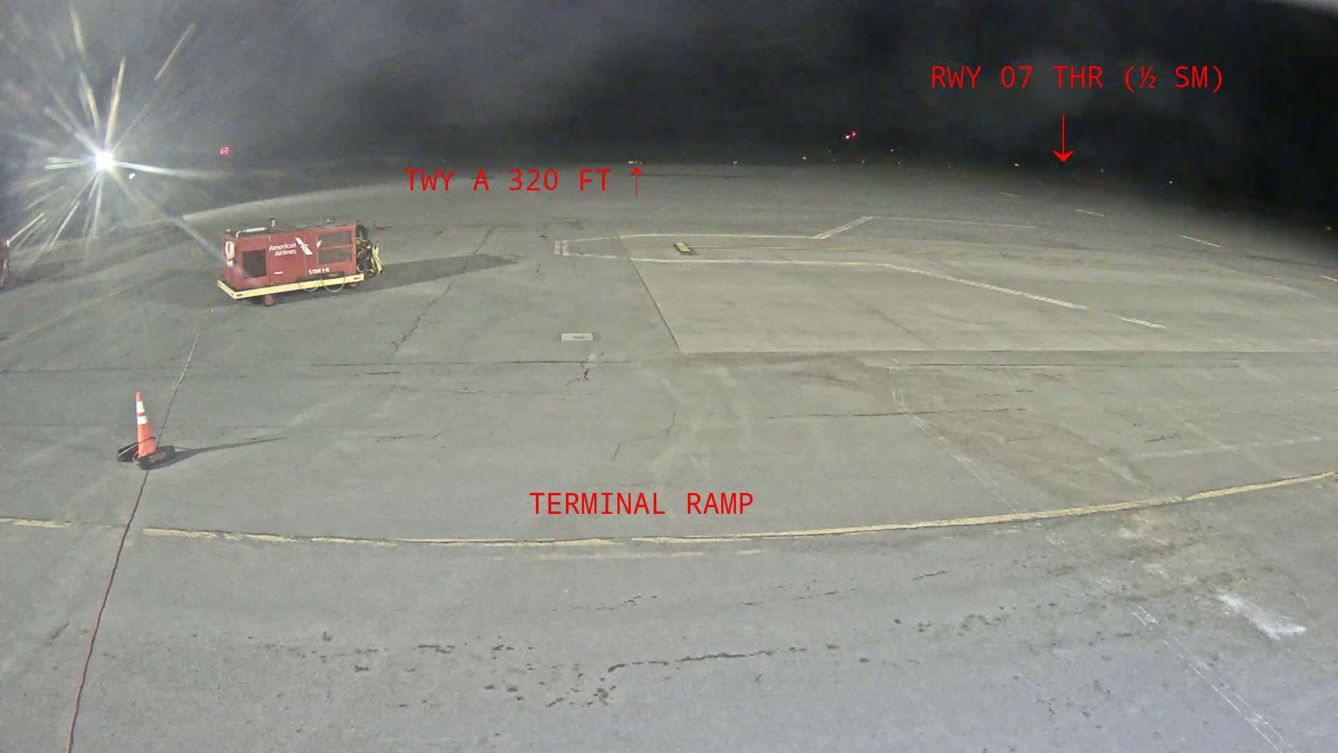A nighttime view of an airport tarmac shows a terminal ramp, a baggage tug, and a traffic cone under a dark, cloudy sky.