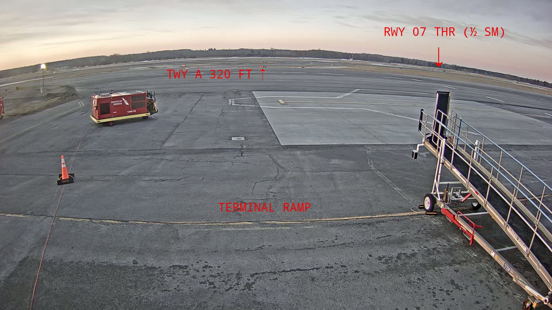 A tarmac at an airport shows a baggage cart, jet bridge, and a runway extending into the distance under a twilight sky.