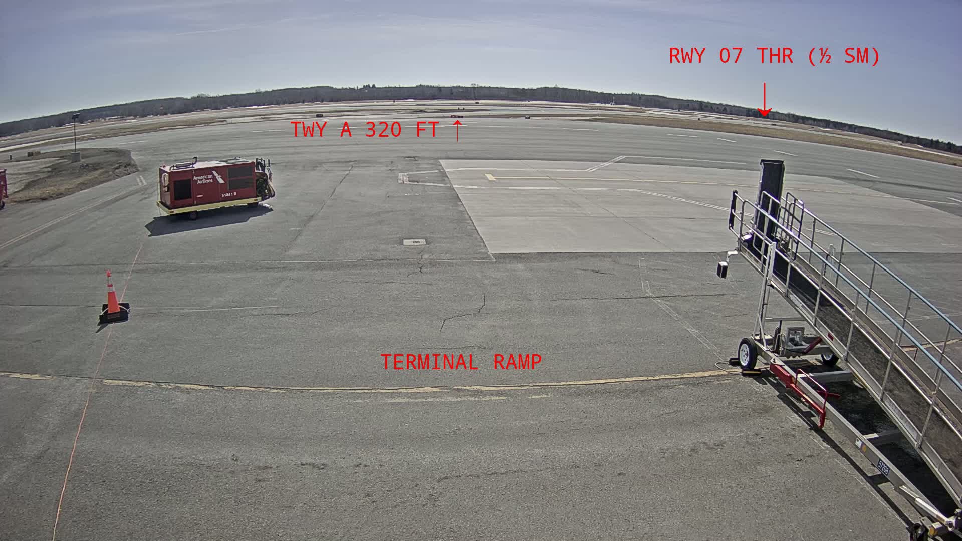 A tarmac with an airport jet bridge, a baggage cart, and a runway visible in the distance under sunny skies.