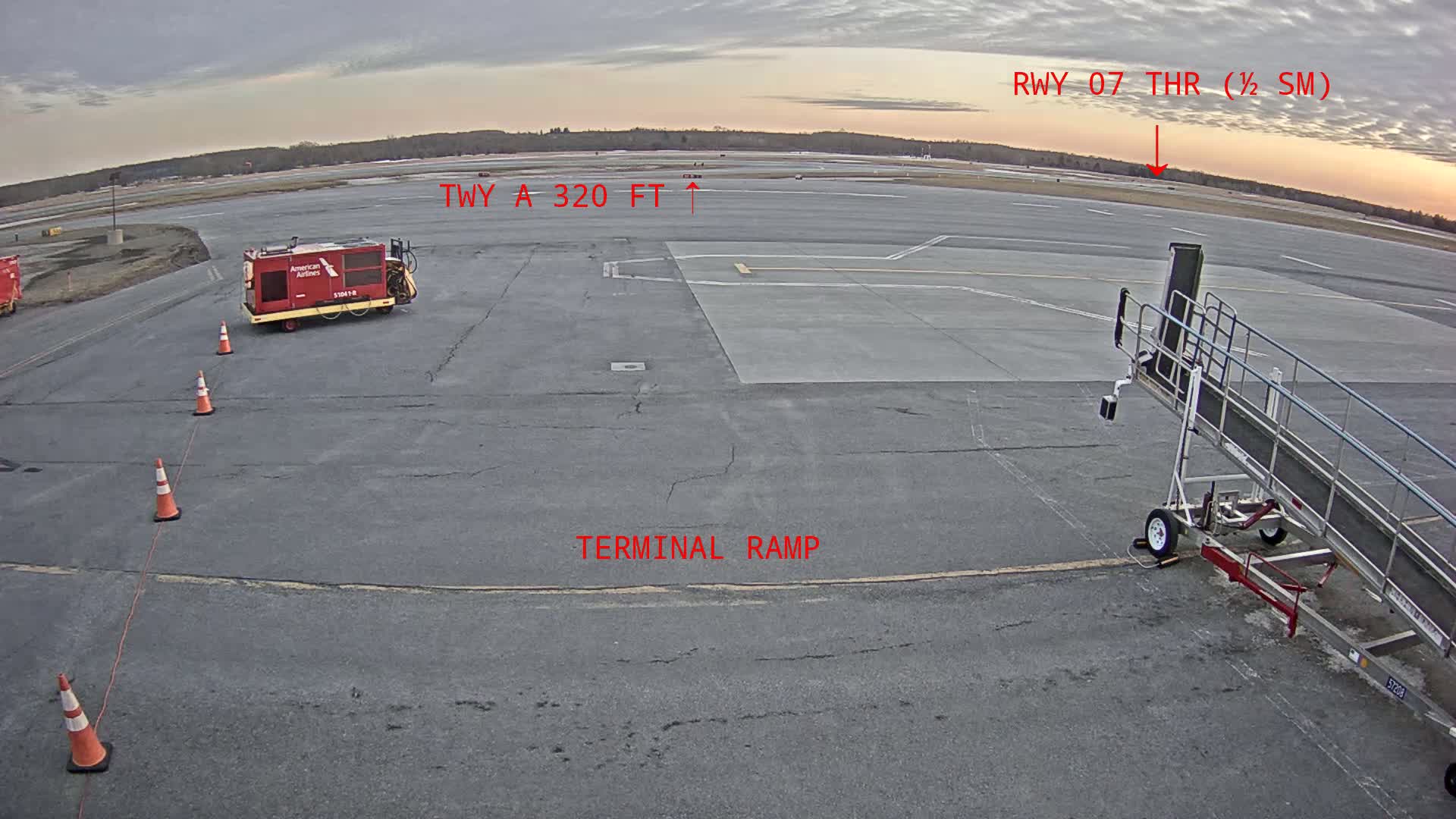An airport tarmac with a jet bridge, a small service vehicle, and orange traffic cones under an overcast sky.