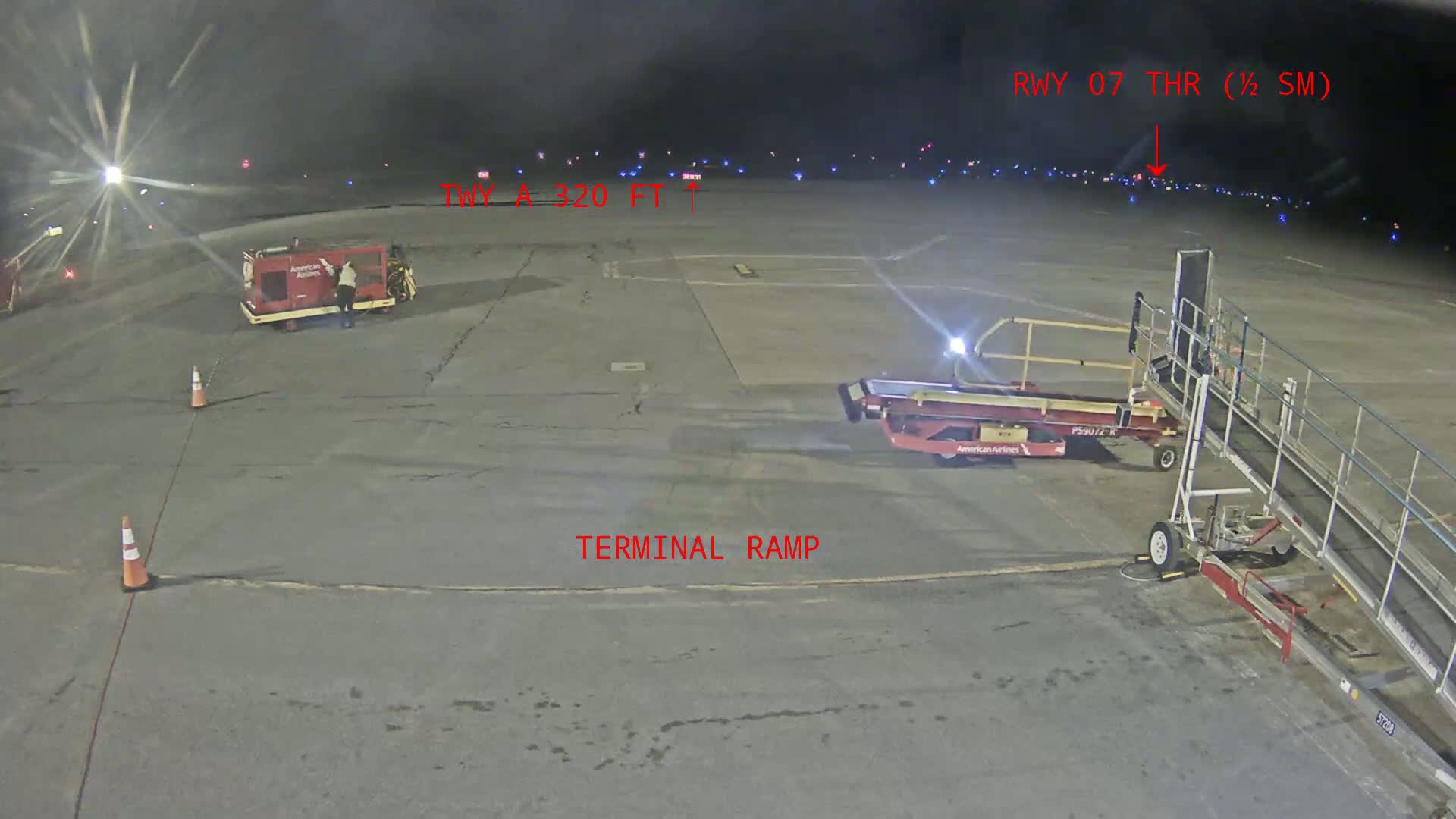 A nighttime airport tarmac scene shows an empty terminal ramp with several pieces of ground support equipment, including a passenger jet bridge, and a distant runway with lights visible.