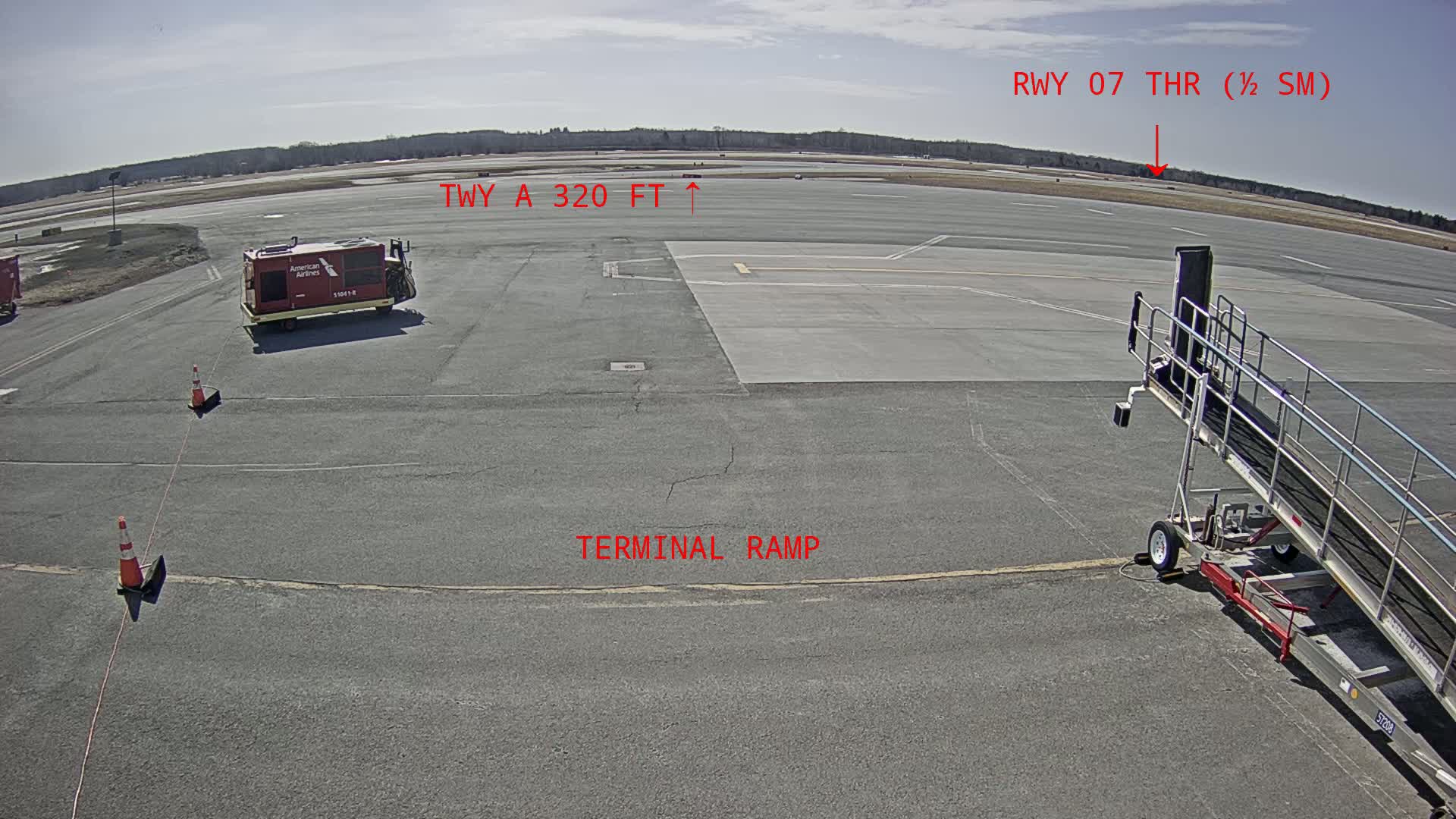 A tarmac under a sunny sky shows an airport terminal ramp, a baggage cart, a jet bridge, and a runway in the distance.