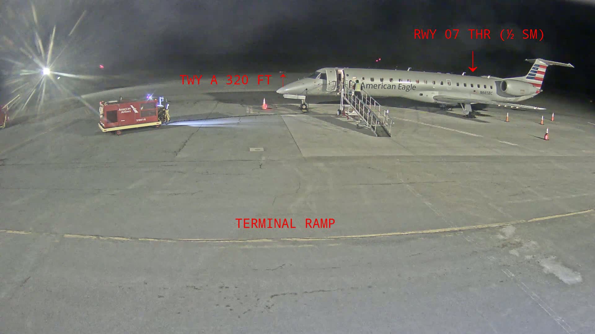 At night, a small American Eagle plane is parked at a terminal ramp, with a baggage cart nearby.