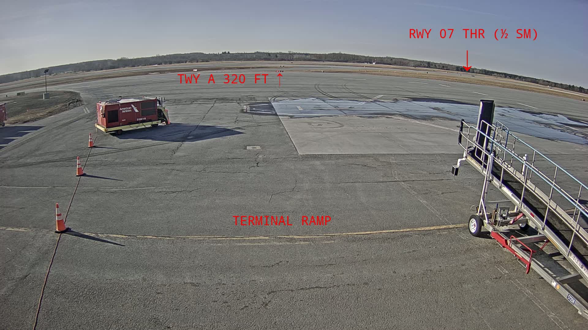 A tarmac under a sunny sky shows an American Airlines baggage cart, a jet bridge, and orange traffic cones near a terminal ramp with a runway in the distance.