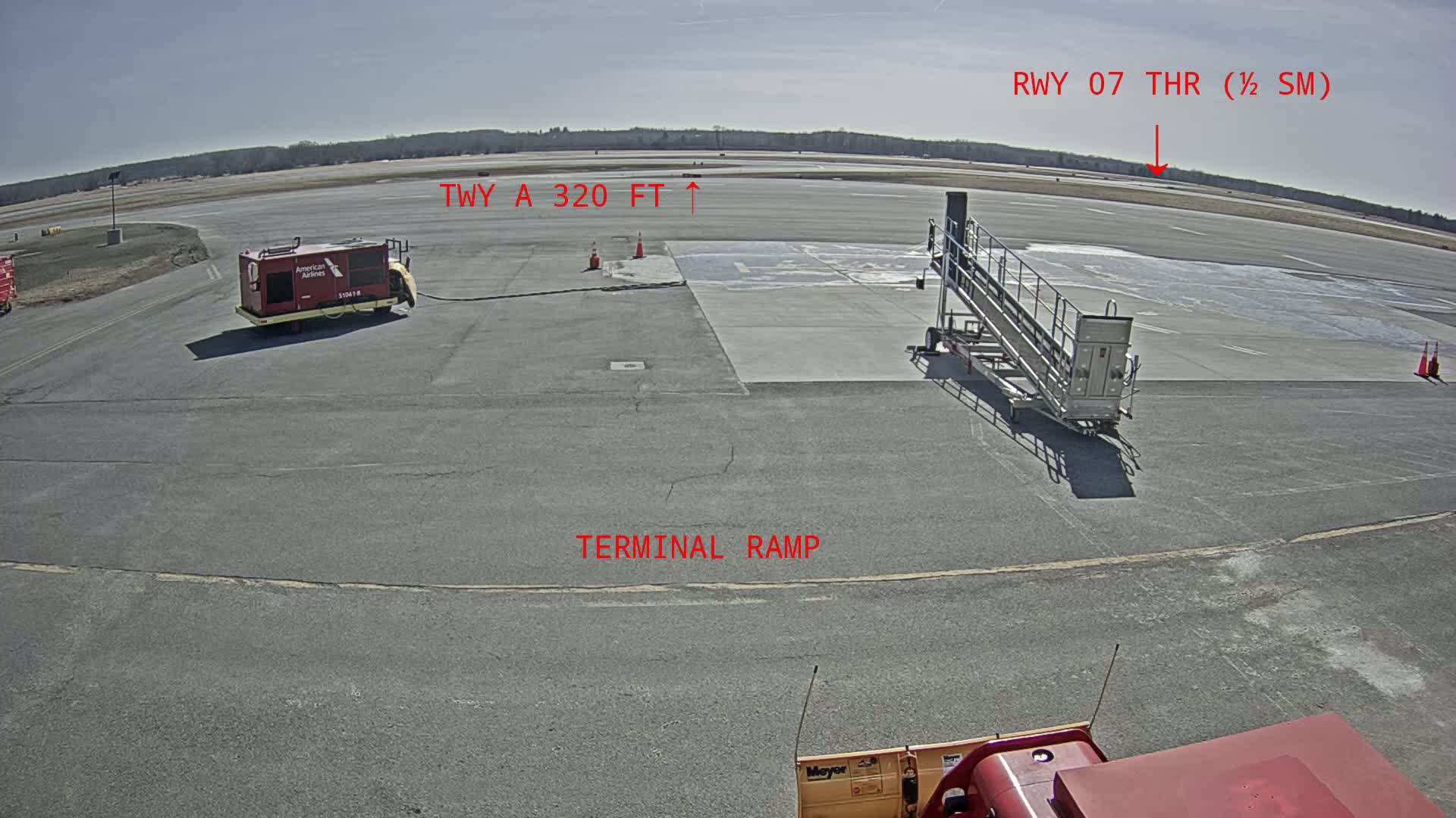 An airport tarmac on a sunny day shows a red ground power unit, a passenger boarding bridge, and part of a snow plow, with a runway visible in the distance.
