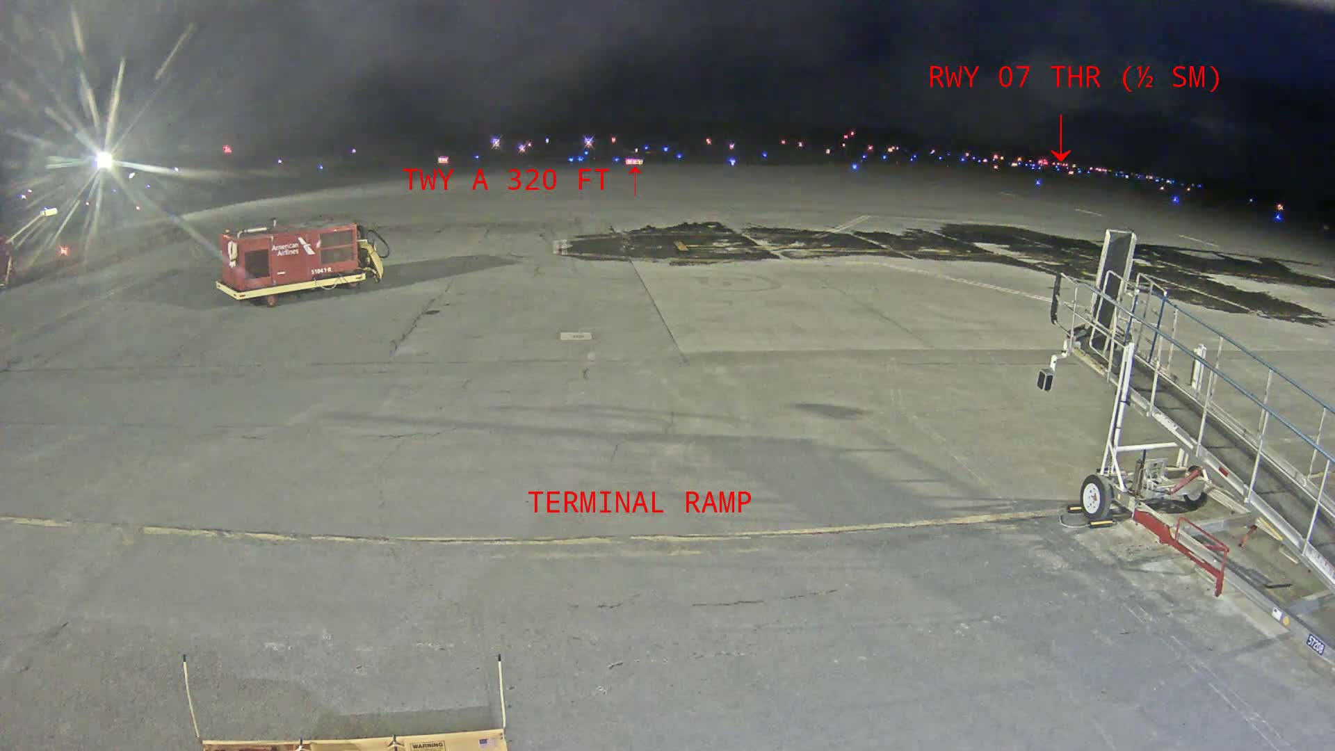 A nighttime view of an airport terminal ramp with a baggage tug and jet bridge, showing a runway in the distance under dark skies.