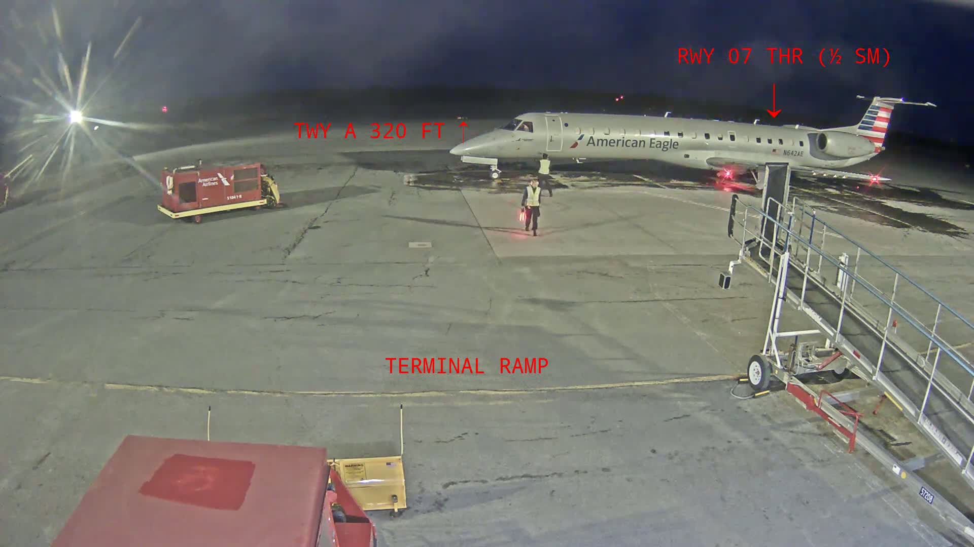 At night, a person walks on an airport tarmac near a parked American Eagle airplane and a jet bridge, with ground support equipment nearby.
