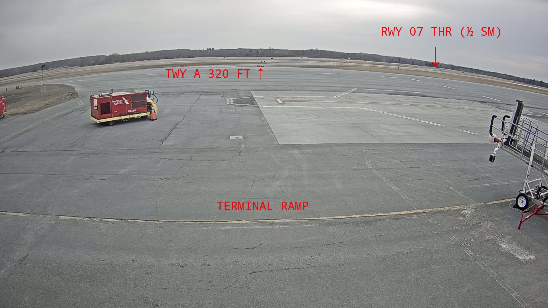 A tarmac view of an airport terminal ramp on an overcast day, showing a baggage tug and an aircraft stairway lift.