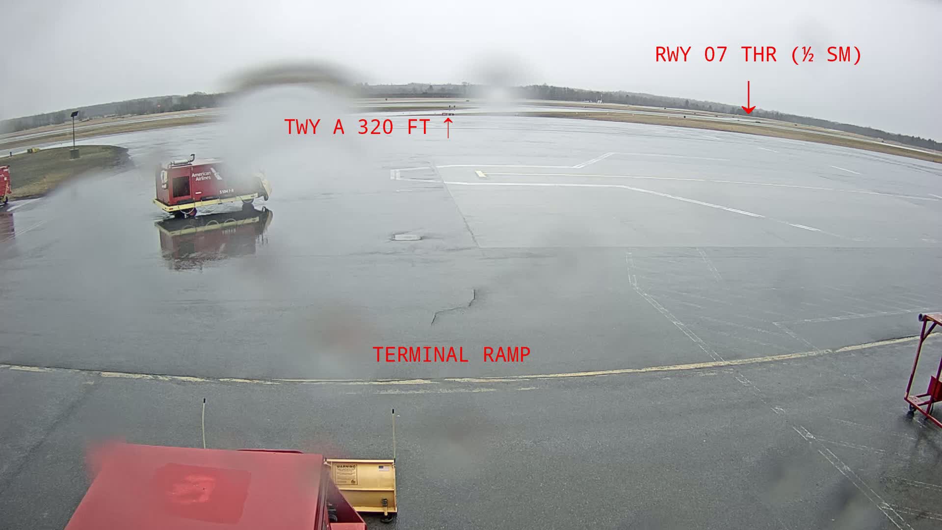 A wet airport tarmac is visible, showing an American Airlines service vehicle and a snow plow in the foreground, with a runway and taxiway in the background under an overcast sky.