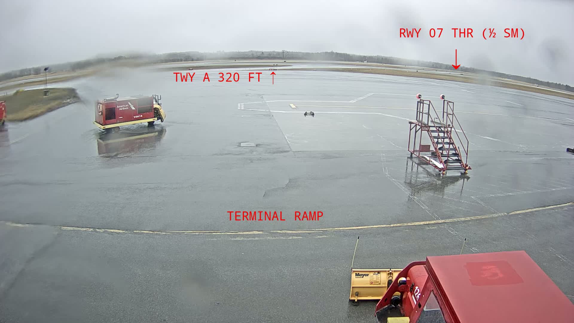 A rain-slicked airport tarmac shows a baggage cart, a snowplow, and a staircase leading to a plane, with a distant runway visible under an overcast sky.