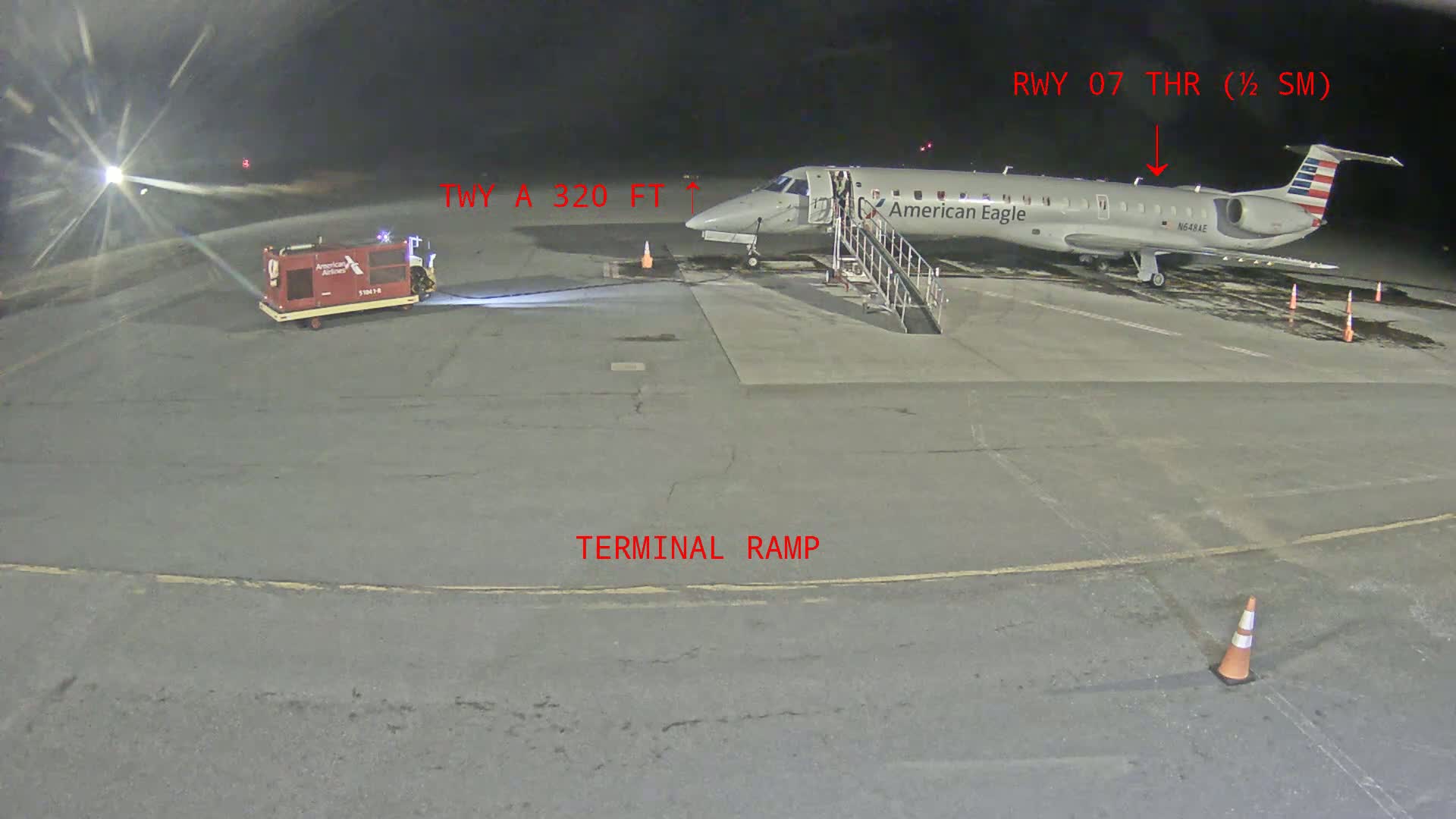 At night, an American Eagle airplane is parked at a terminal ramp, with a baggage tug nearby.
