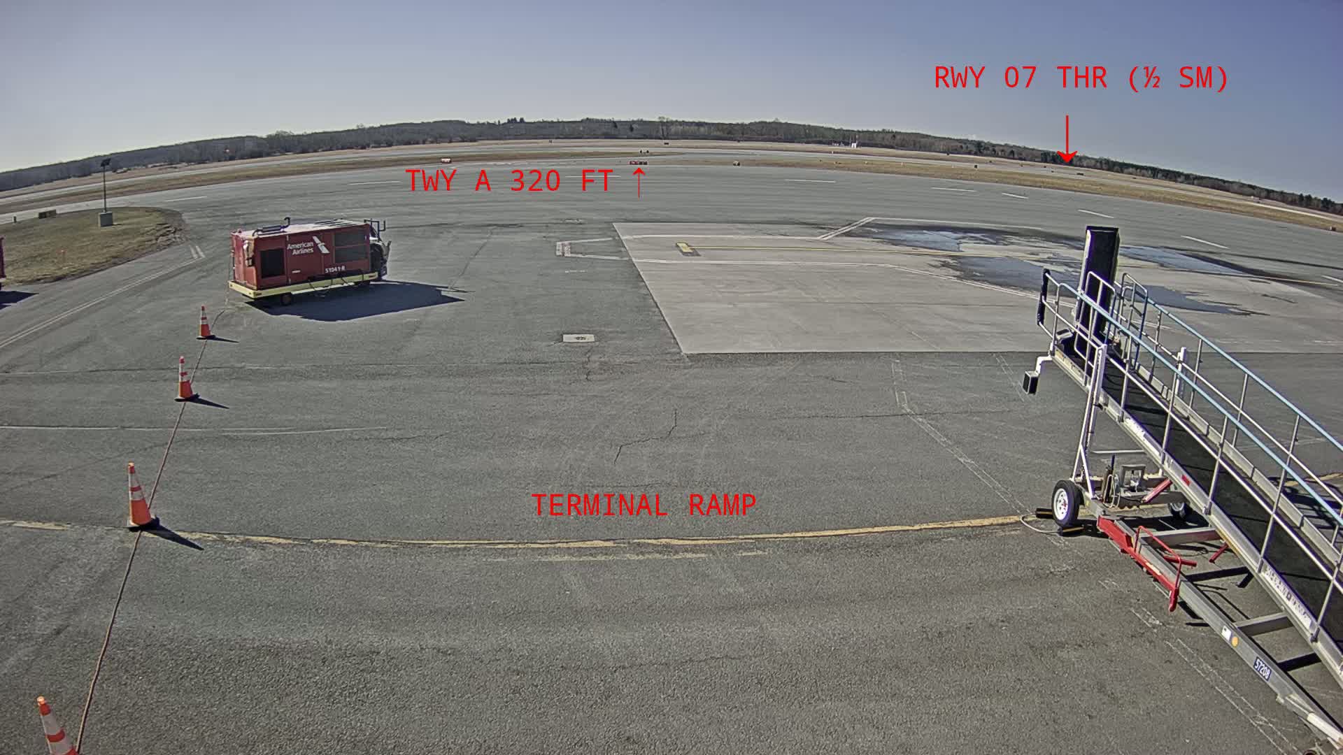 A tarmac with an American Airlines baggage cart, jet bridge, and orange traffic cones under a clear, sunny sky.