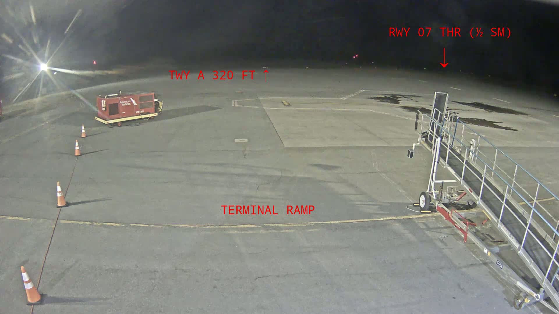 A nighttime view of an airport tarmac shows a jet bridge, ground power unit, and several orange traffic cones under dark skies.