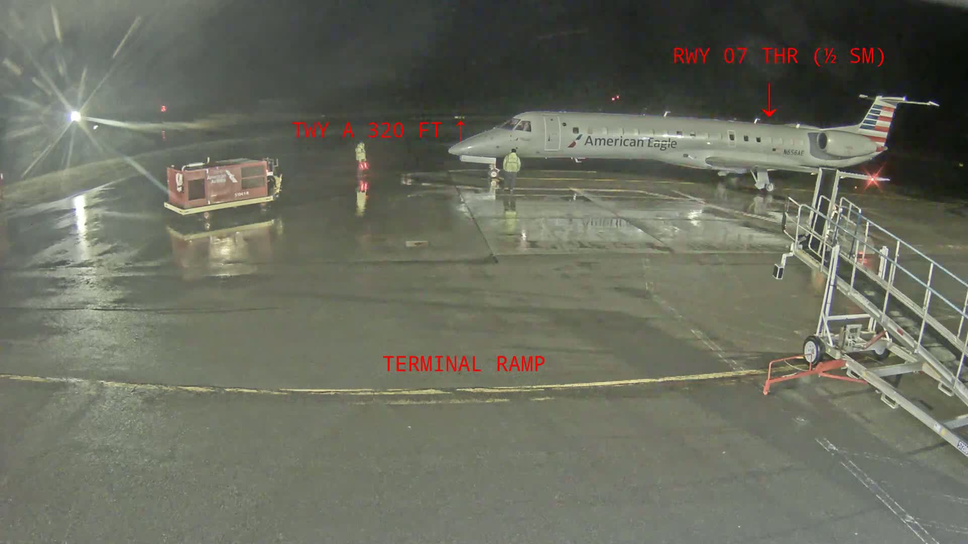 A wet tarmac at night shows an American Eagle plane parked at the terminal with ground support equipment present.