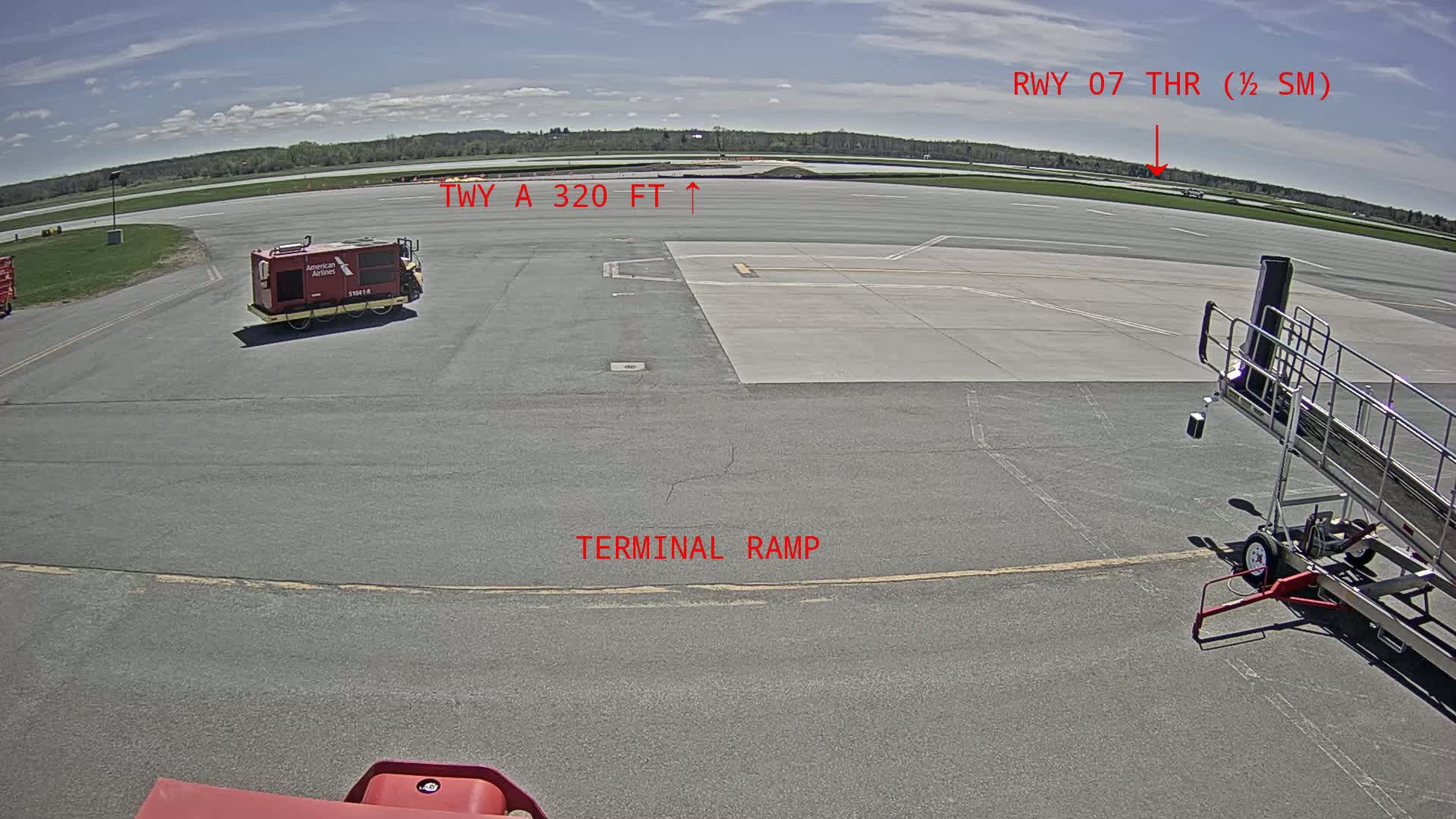 A tarmac with a red baggage cart and jet bridge is visible on a sunny day, with a runway and grassy area extending into the background.