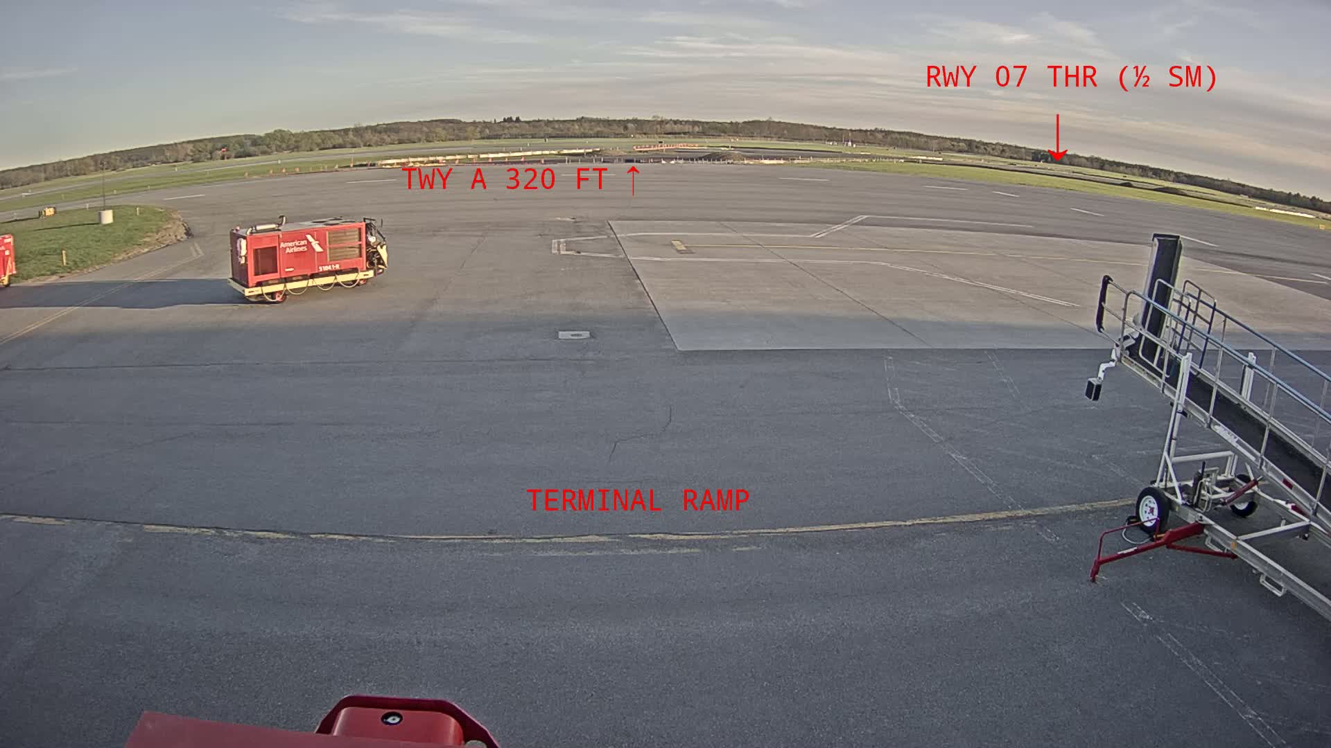 A tarmac at an airport on a partly sunny day shows a red ground support vehicle, a jetway, and a runway in the distance.