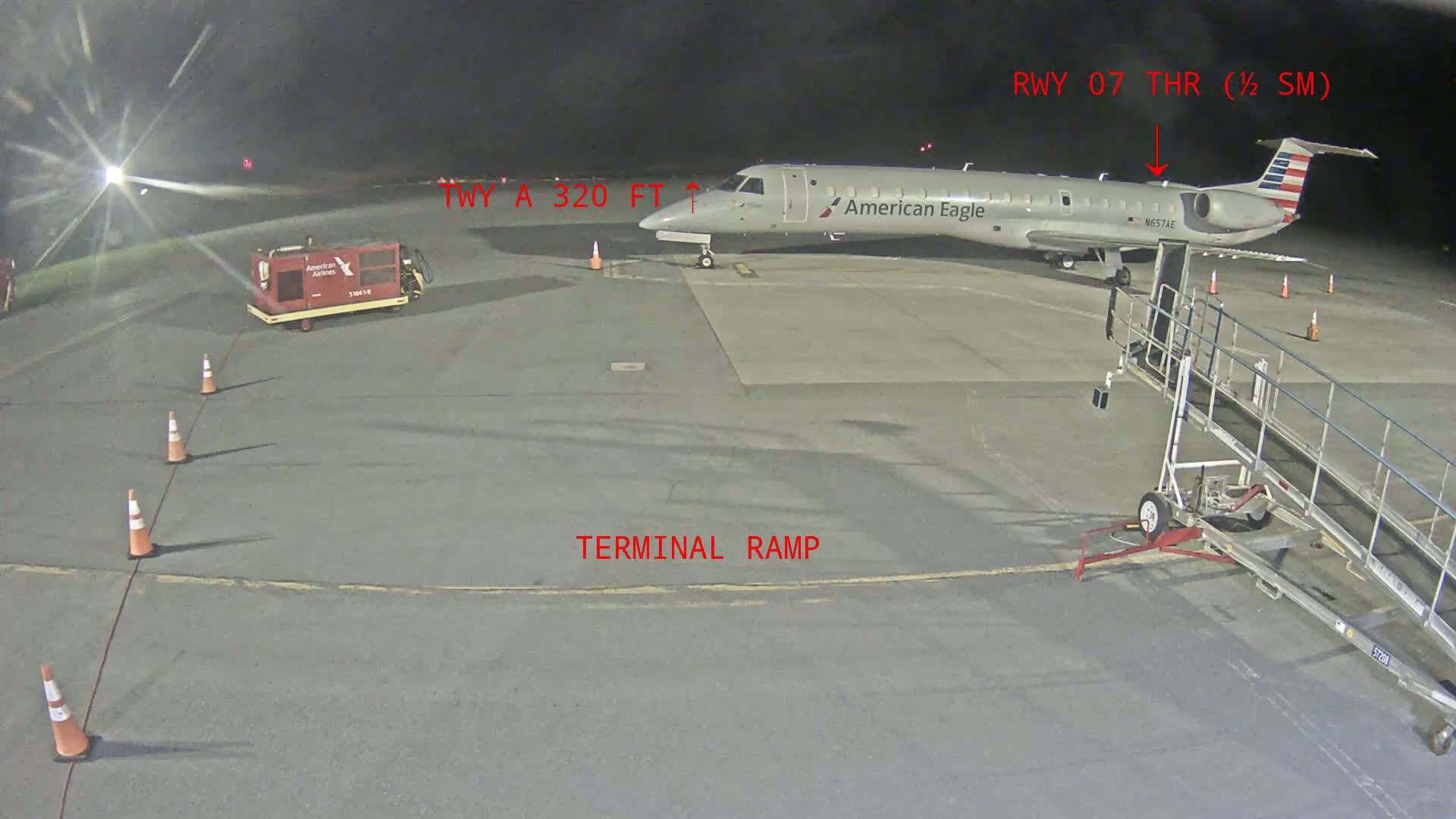At night, a tarmac shows an American Eagle airplane parked next to a jet bridge, with ground support equipment and orange cones nearby.