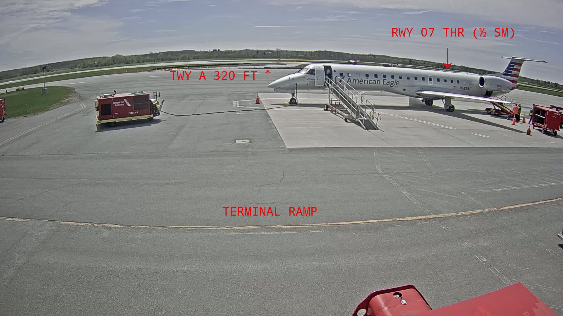 An American Eagle regional jet is parked at a terminal ramp on a sunny day at an airport, with ground support equipment nearby.