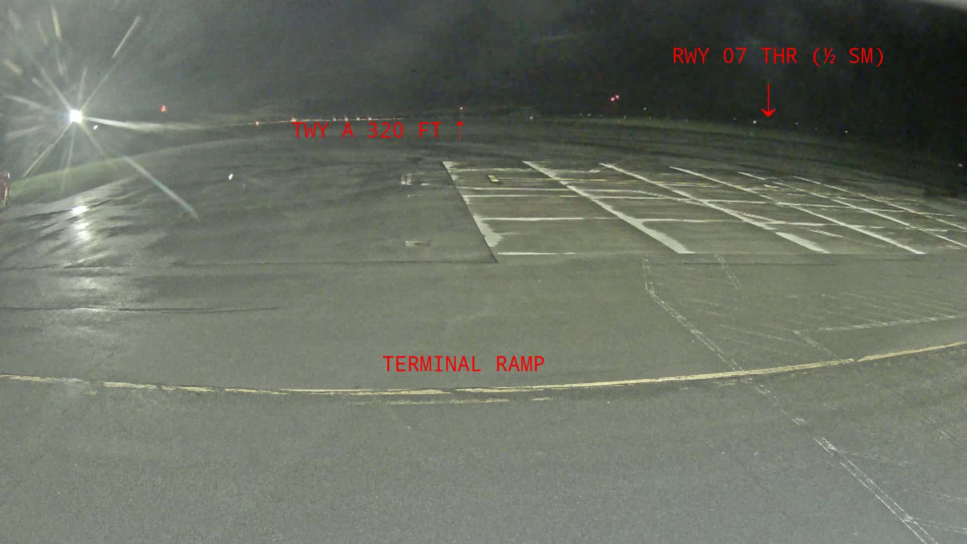 A wet, dark asphalt tarmac area at night shows a terminal ramp and marked parking spaces, with a runway visible in the distance under dark, nighttime conditions.