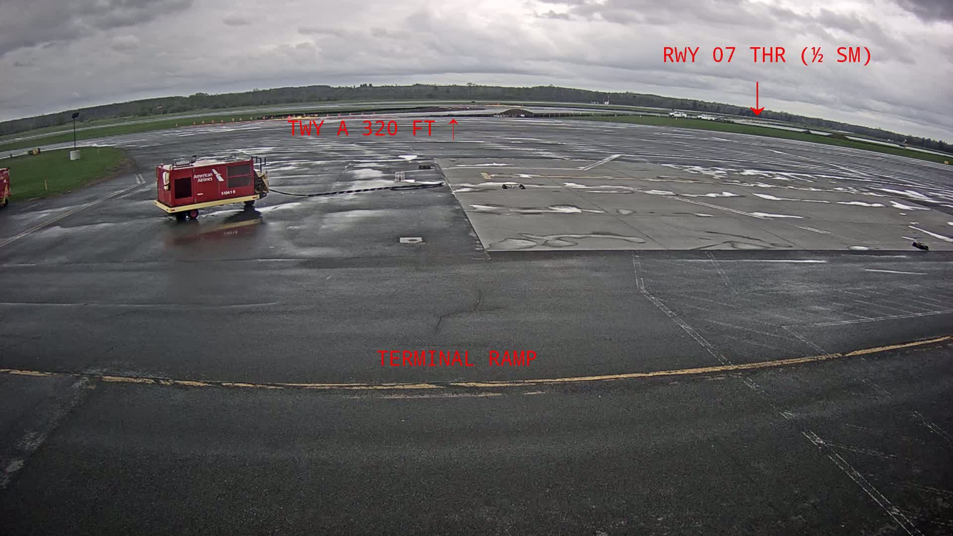 A wet airport tarmac under an overcast sky shows a small red ground support vehicle and a distant view of a runway.