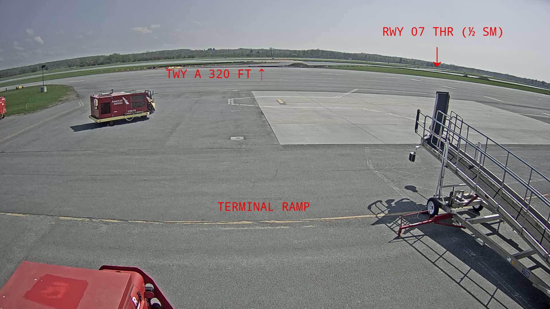 A tarmac with an airport jet bridge, ground support equipment, and a runway visible in the distance under sunny skies.