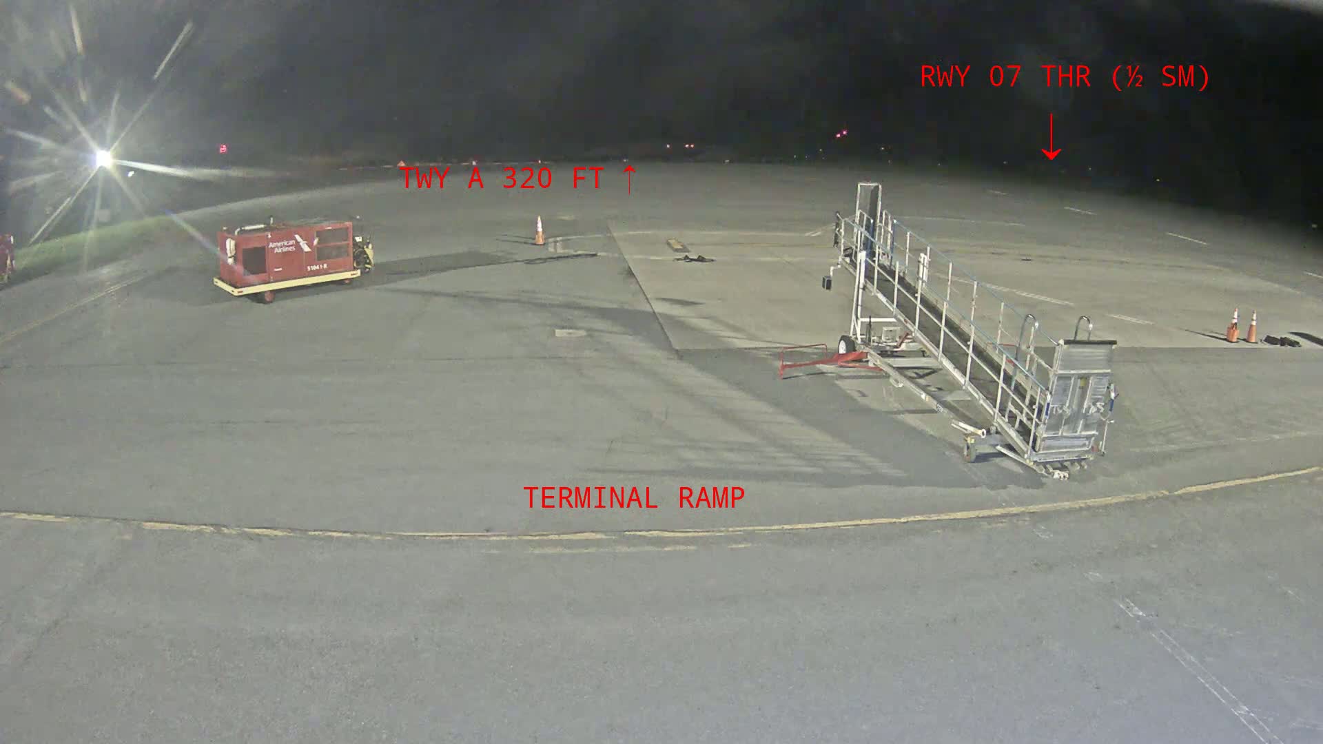 A nighttime airport tarmac shows a baggage cart, a passenger boarding bridge, and runway markings.