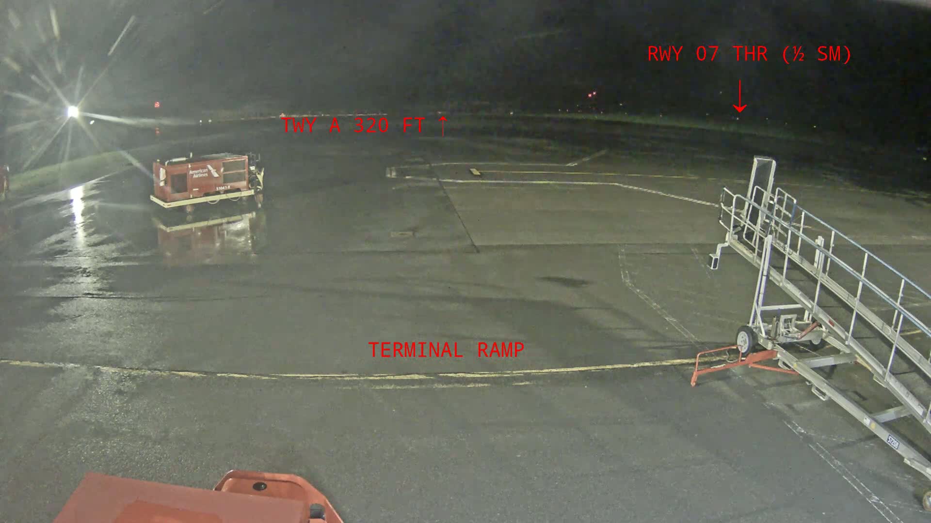 A wet airport tarmac at night shows a baggage cart, a jet bridge, and a service vehicle.
