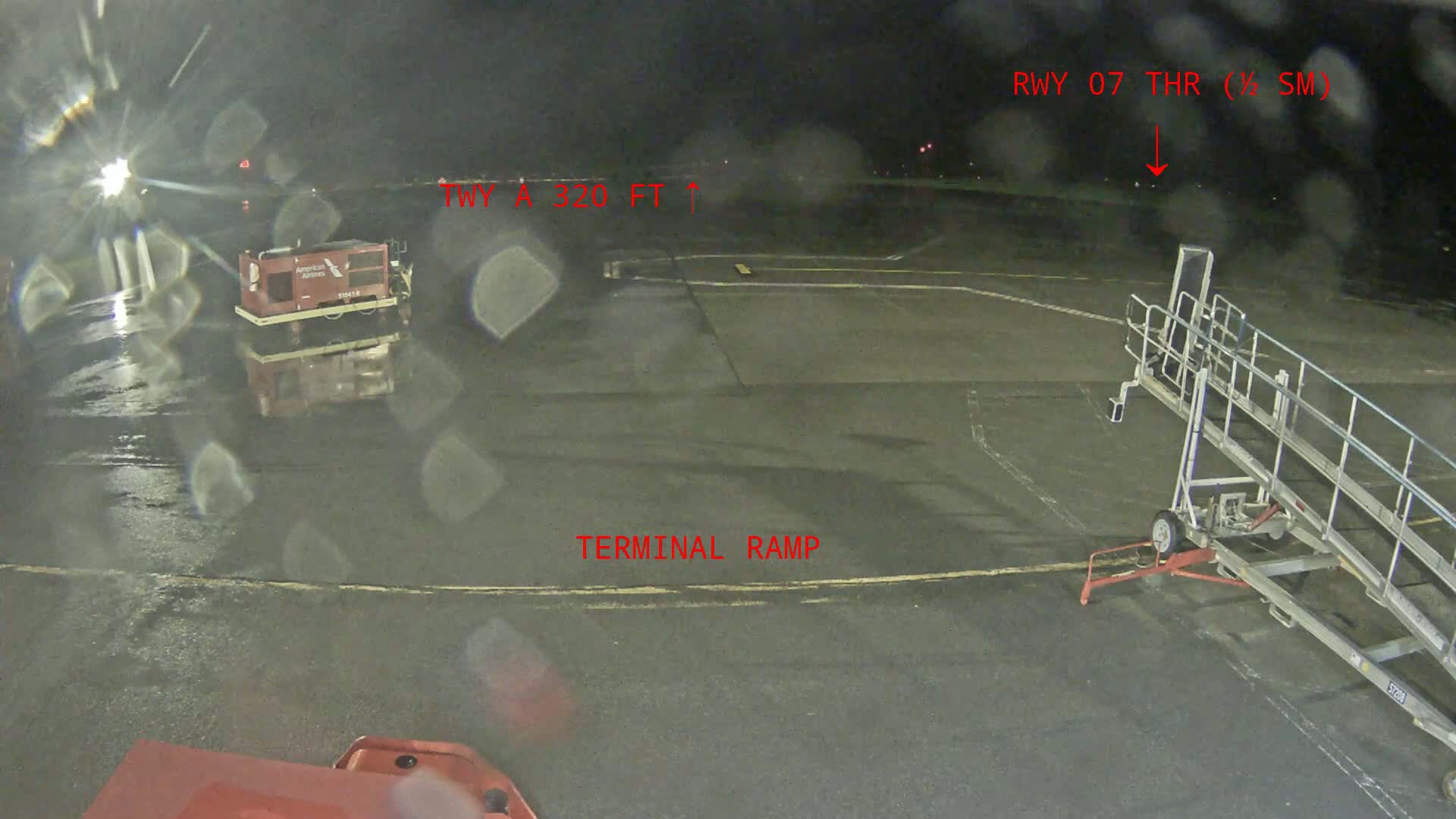 A wet airport tarmac at night shows a baggage cart, an aircraft jet bridge, and faint lights in the distance.