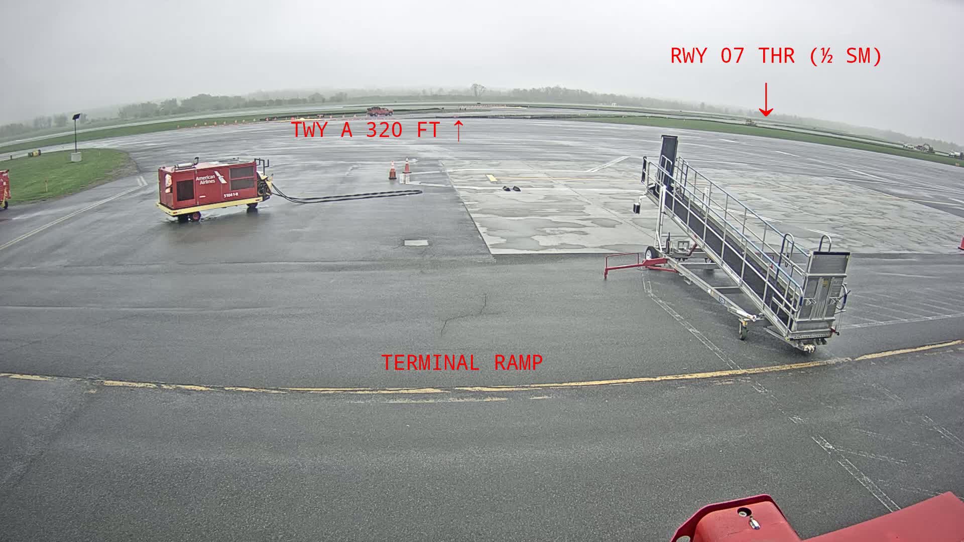 A wet airport tarmac shows a baggage tug, a passenger boarding ramp, and a runway in the distance under an overcast sky.