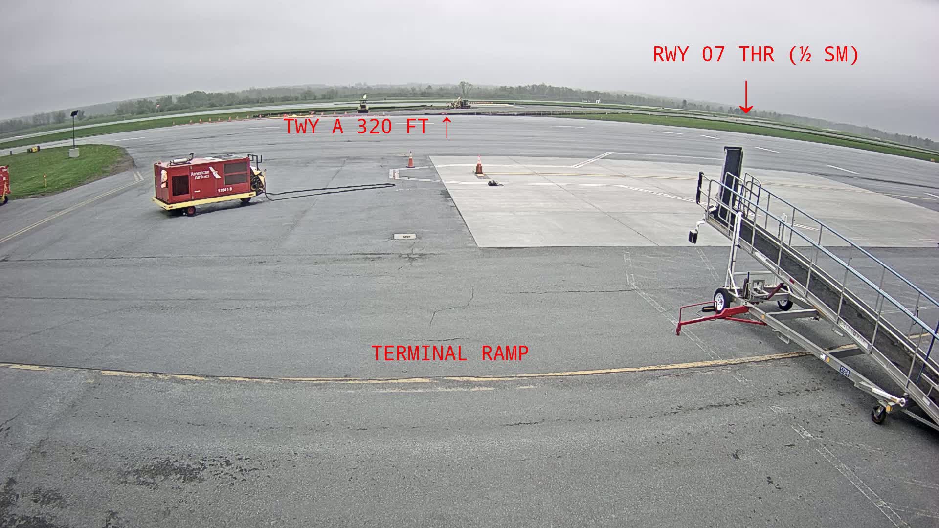 An overcast day at an airport shows a tarmac with a jet bridge, ground support equipment, and a runway in the distance.