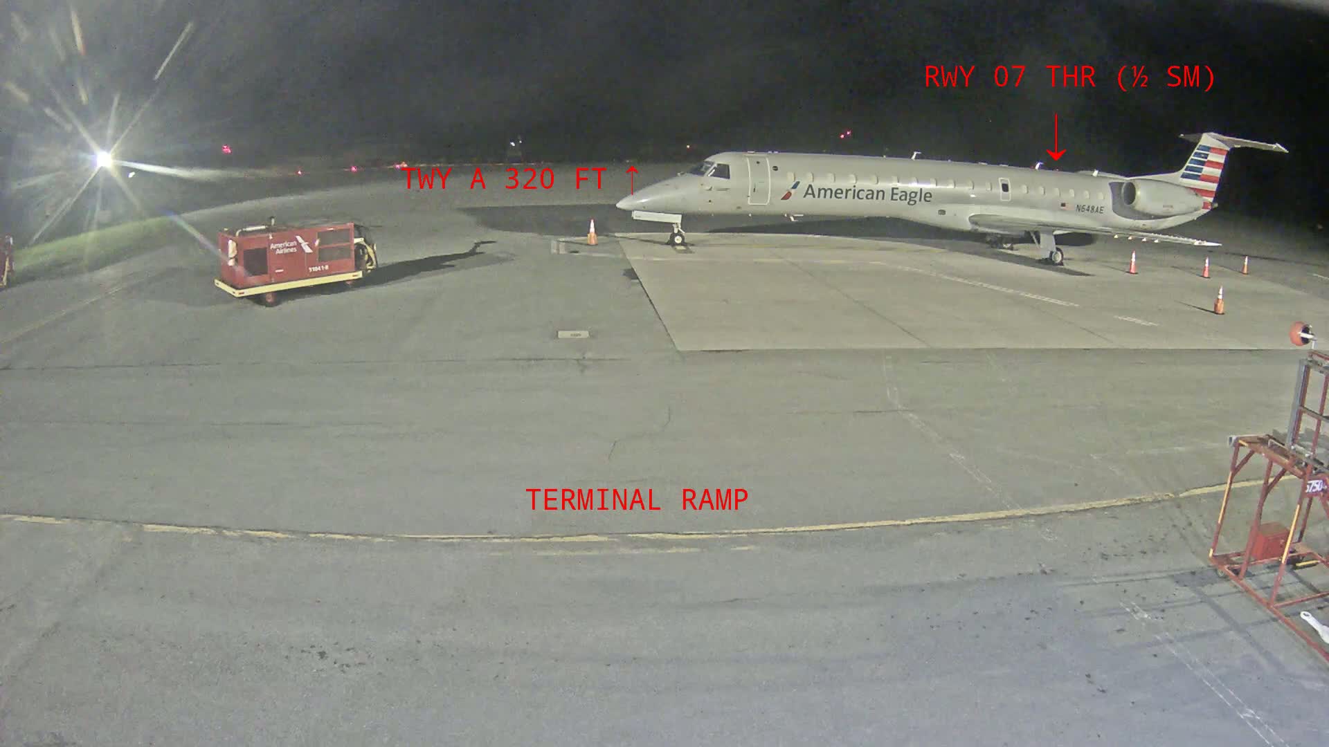 At night, a small American Eagle plane is parked on a terminal ramp at an airport, near a baggage cart.