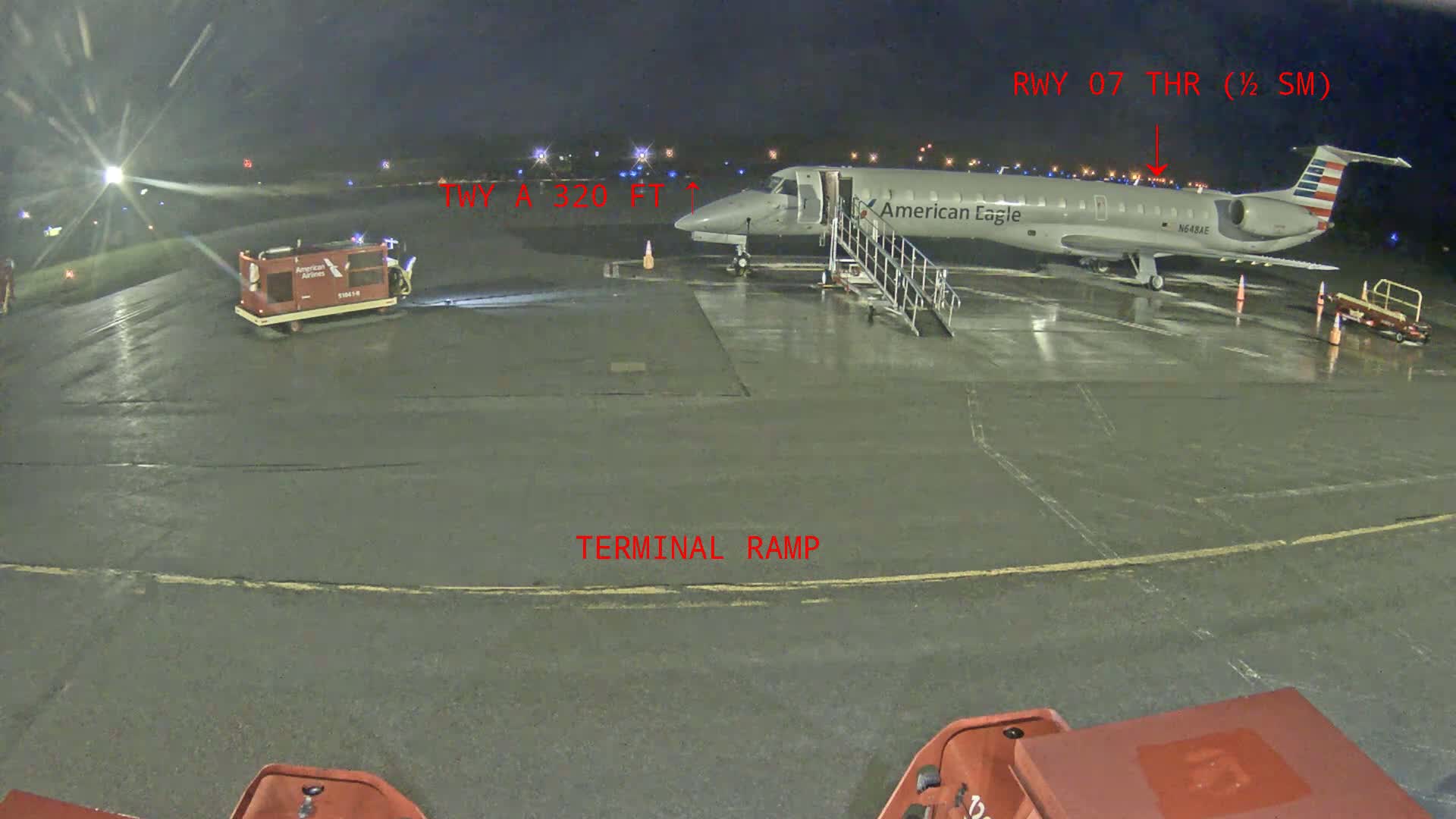 At night, a wet American Eagle regional jet is parked at a terminal ramp with ground support equipment nearby.